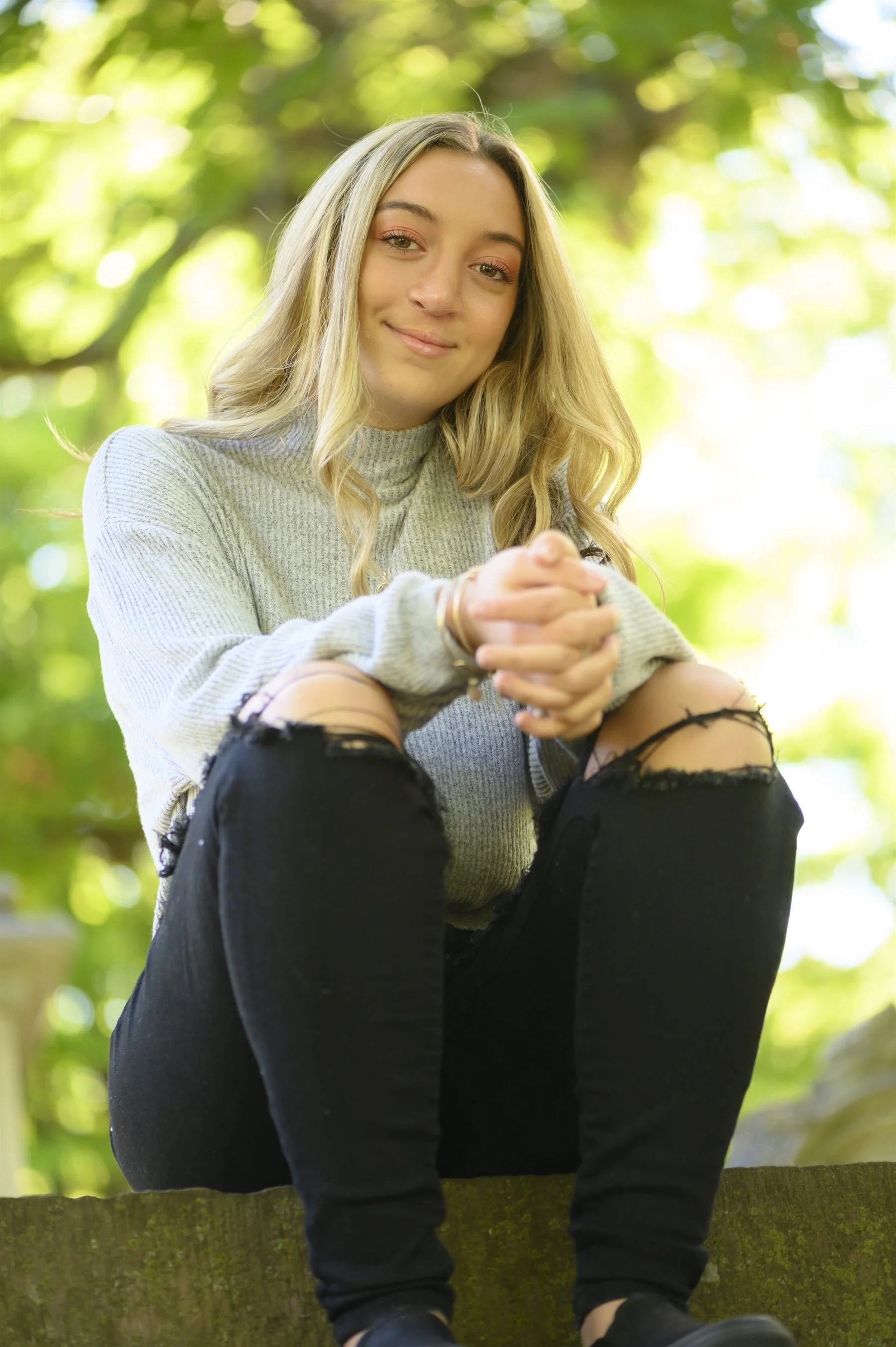 Young woman sitting outdoors, wearing a gray sweater and black ripped jeans, surrounded by greenery.