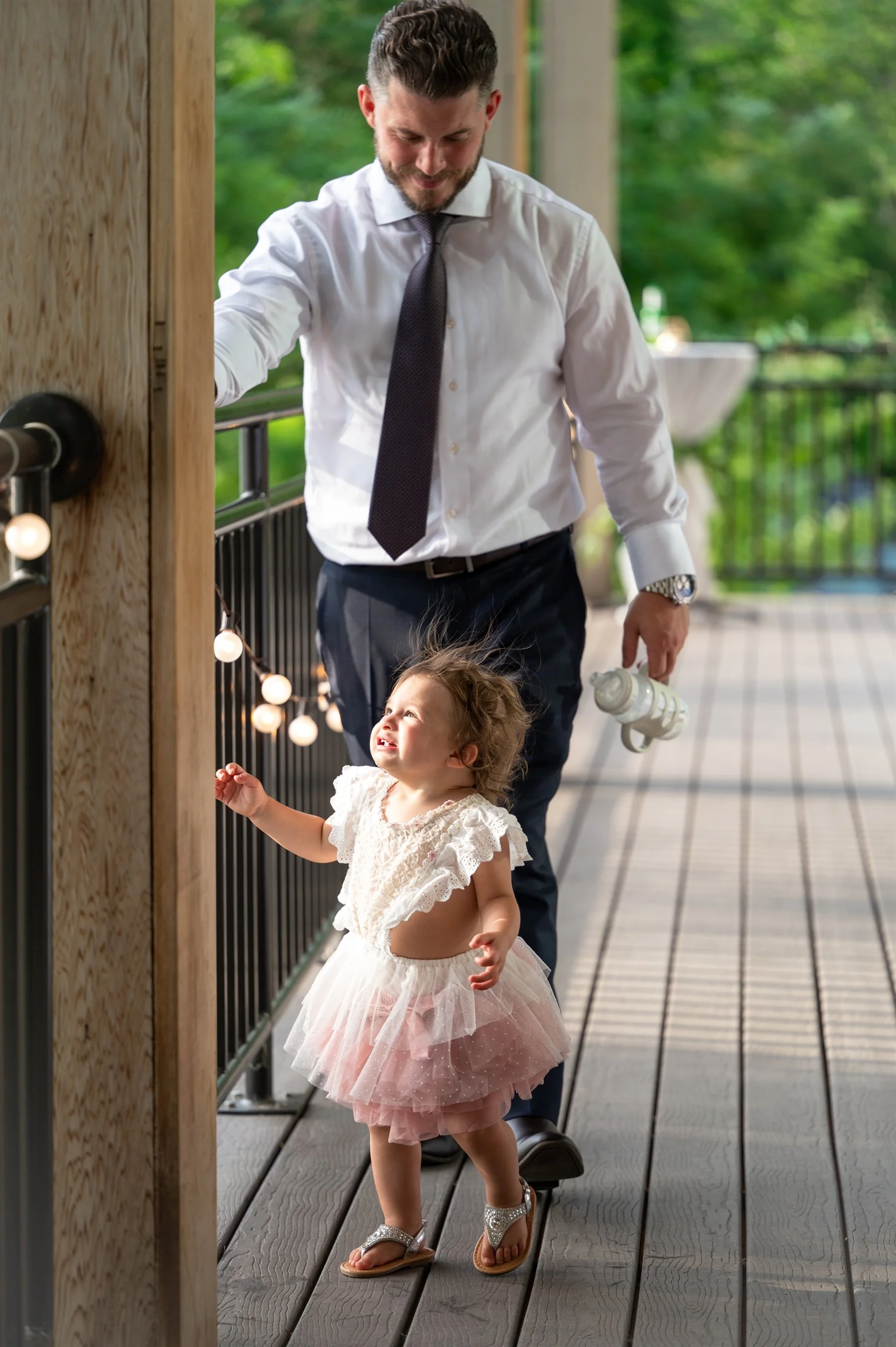 A man in a white shirt and tie stands on a wooden deck, smiling and holding a drink container. A young girl in a pink tutu dress and sandals looks up while touching string lights along the railing. The setting is outdoors with greenery in the backgro