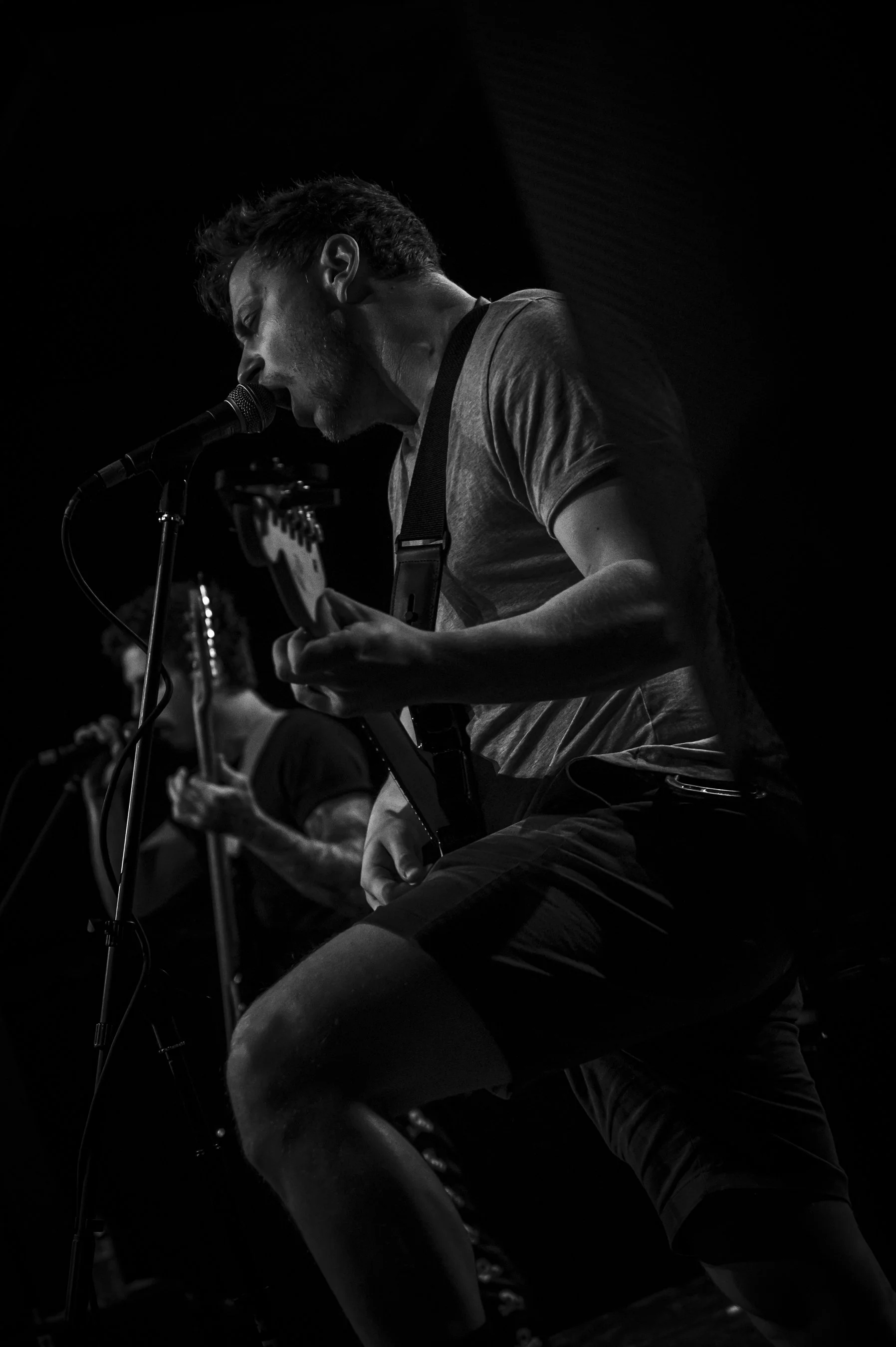 Black and white photo of a man playing electric guitar and singing into a microphone during a live performance.