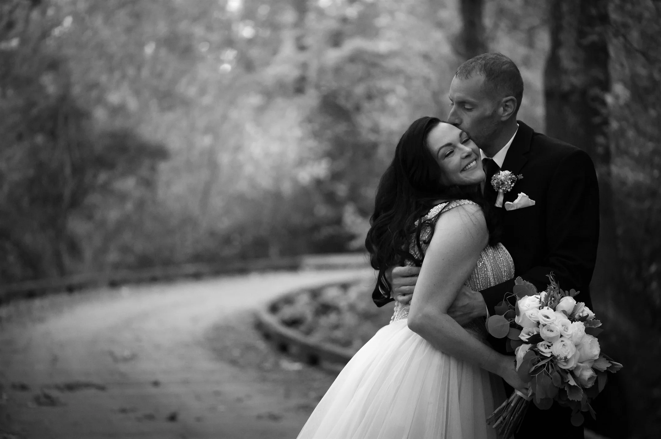 Black and white photo of a bride and groom embracing outdoors, with the groom kissing the bride on the forehead and the bride holding a bouquet of flowers.