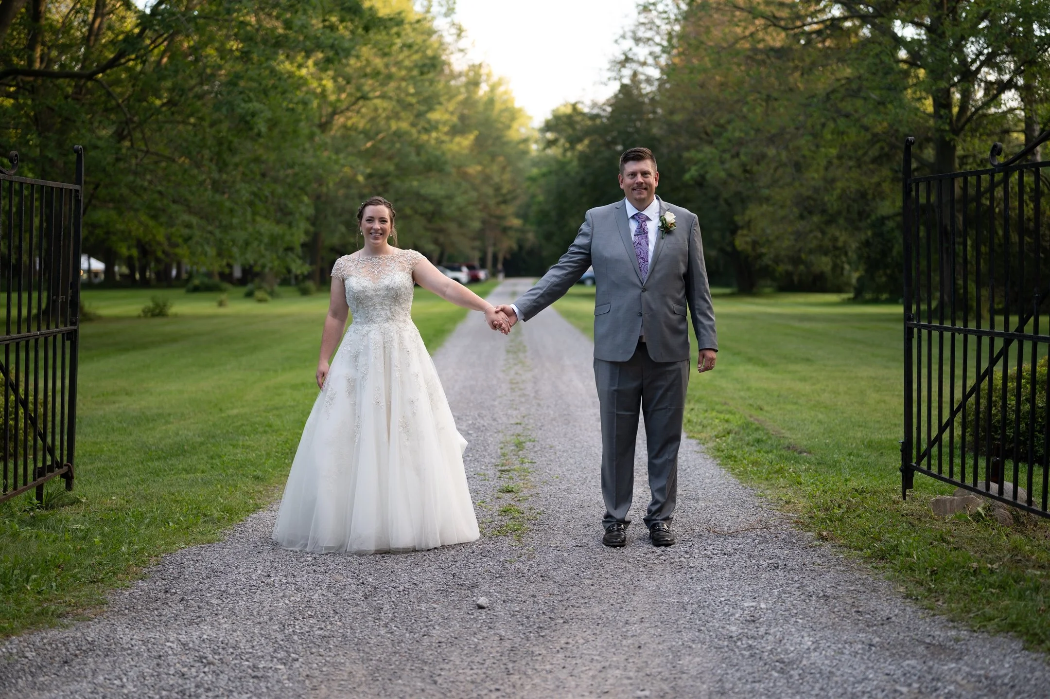 Bride in white wedding dress and groom in gray suit holding hands, standing on a gravel path between open wrought iron gates, surrounded by green grass and trees, with cars in the background.
