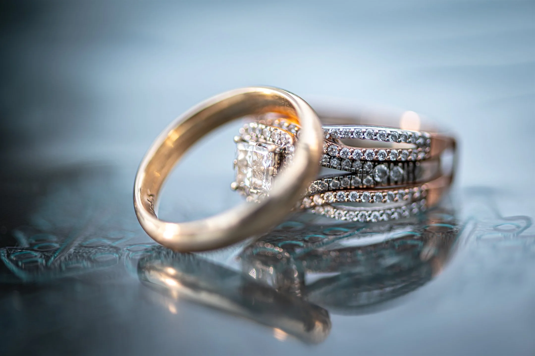 Close-up of a gold wedding band and a diamond-encrusted engagement ring on a reflective surface.