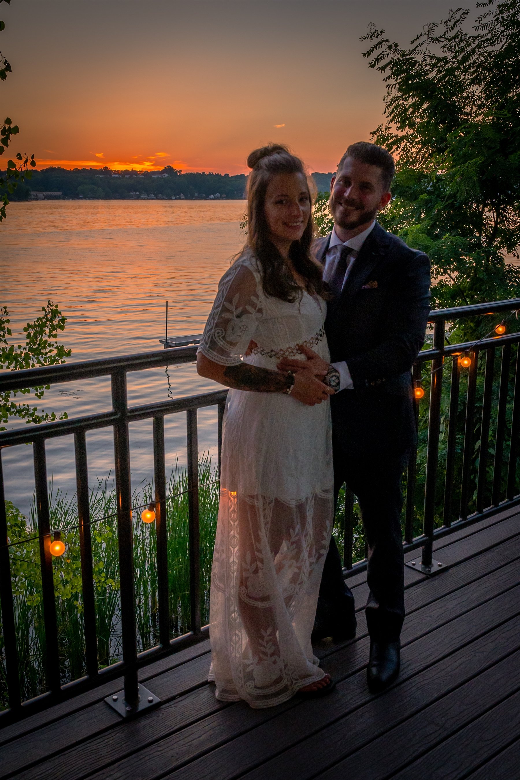 Couple posing on waterfront balcony at sunset, woman in white lace dress and man in dark suit, with string lights and trees in background.