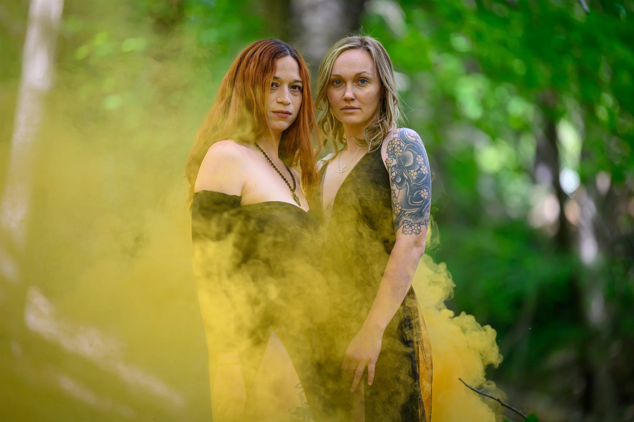 Two women in a forest setting surrounded by yellow smoke. One woman has long red hair and a dark off-shoulder dress, while the other has blonde hair, a tattooed arm, and a similar dress. The background is lush and green.