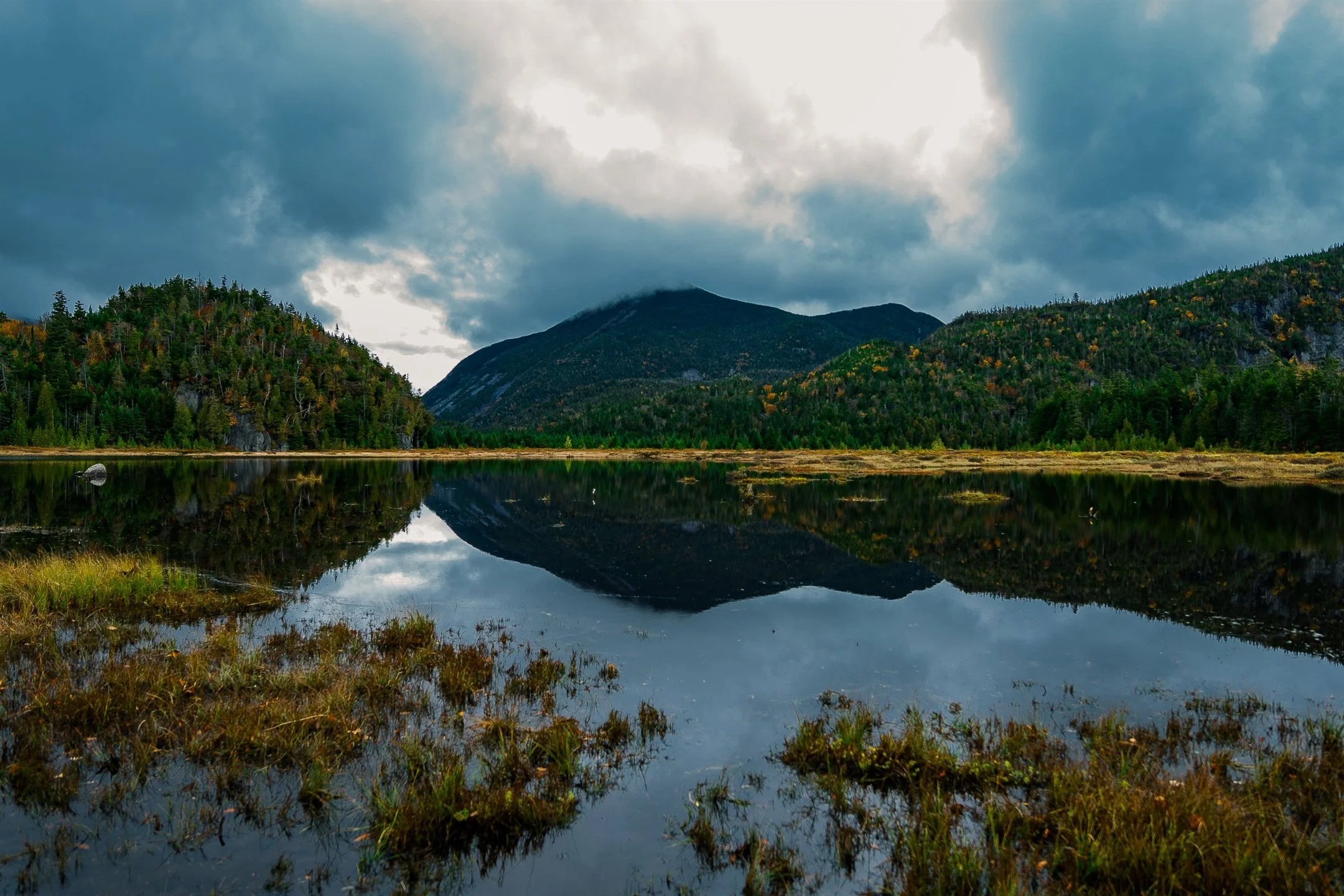 Serene landscape with a calm lake reflecting surrounding lush green mountains and overcast sky.