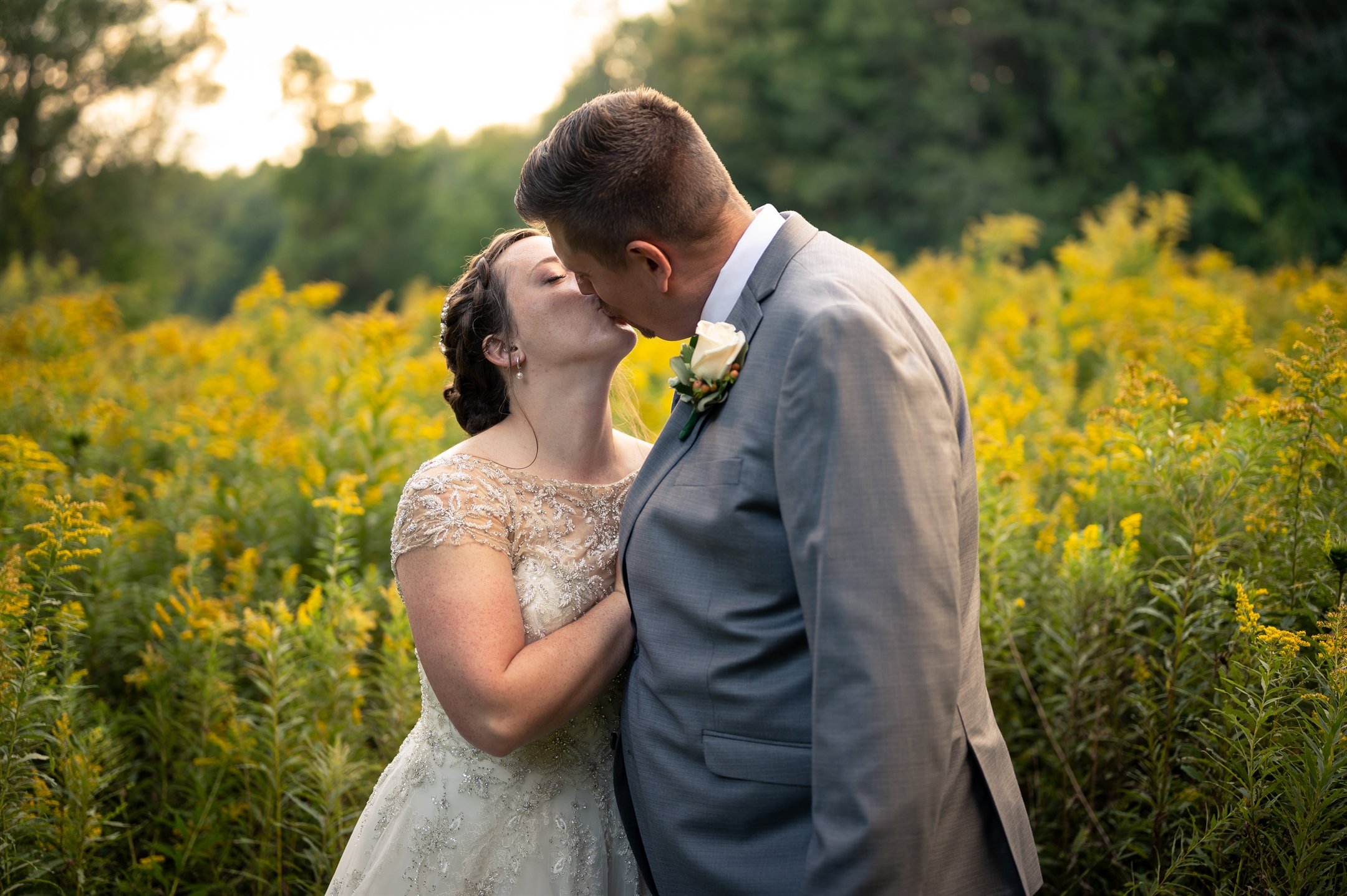 Bride and groom kissing in a field of yellow flowers, wearing wedding attire.