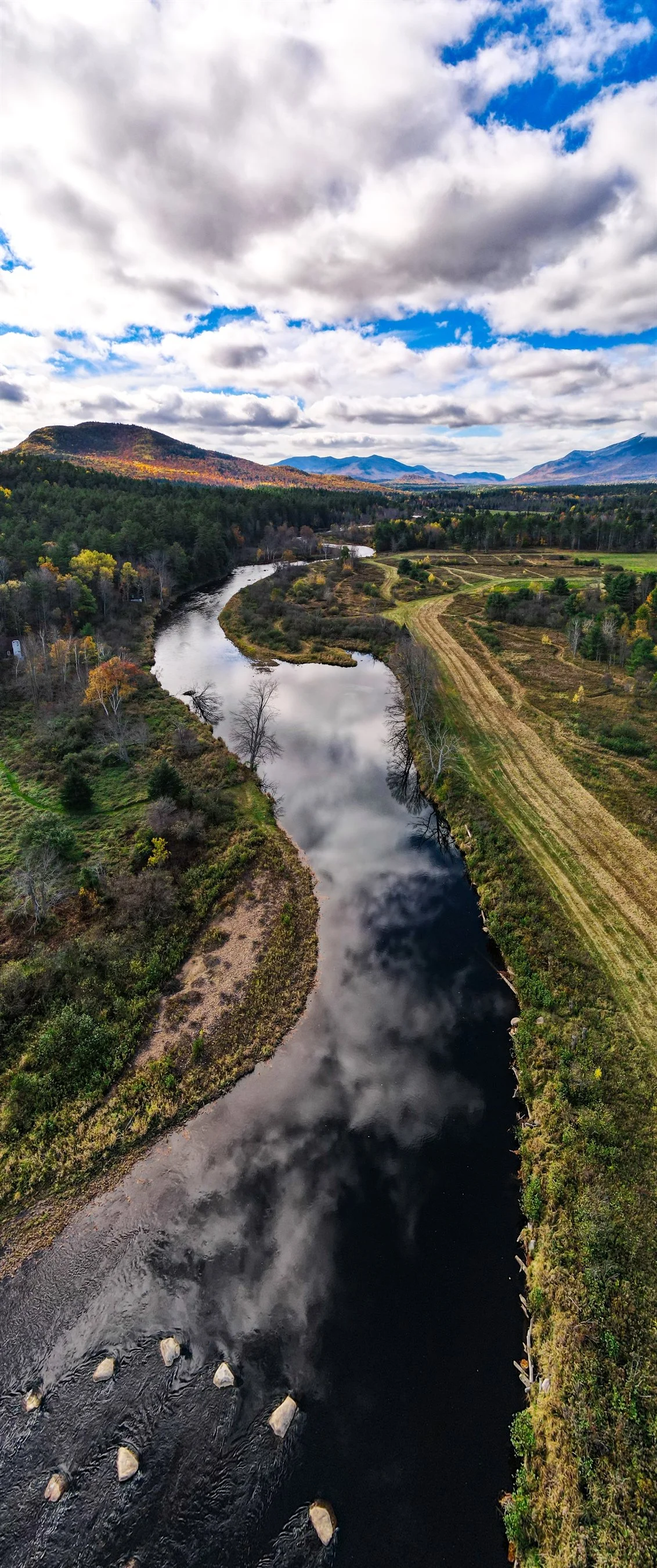 Aerial view of a winding river surrounded by lush greenery, trees, and hills under a cloudy sky.