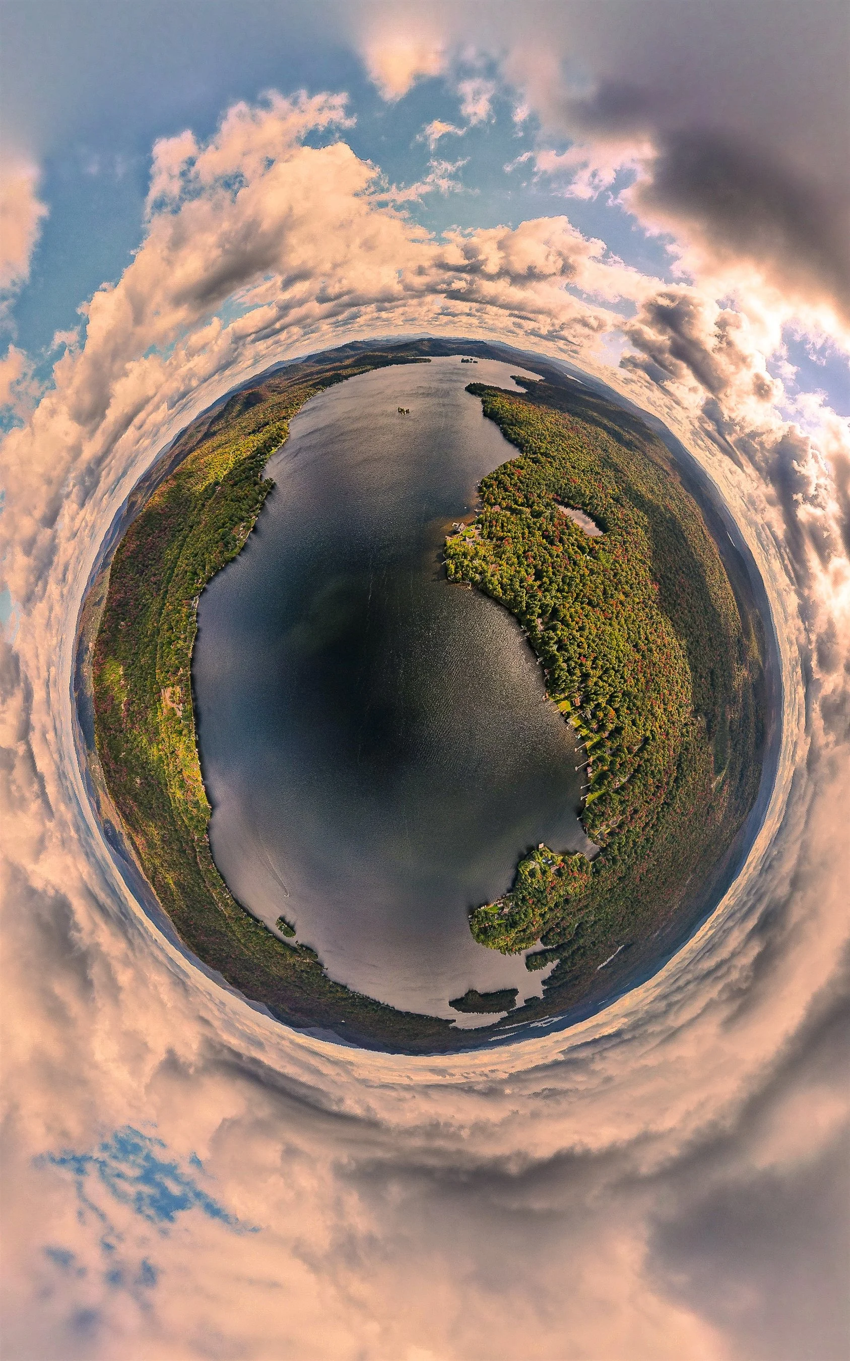 Little planet aerial view of a lake surrounded by forests and mountains, with a cloudy sky.