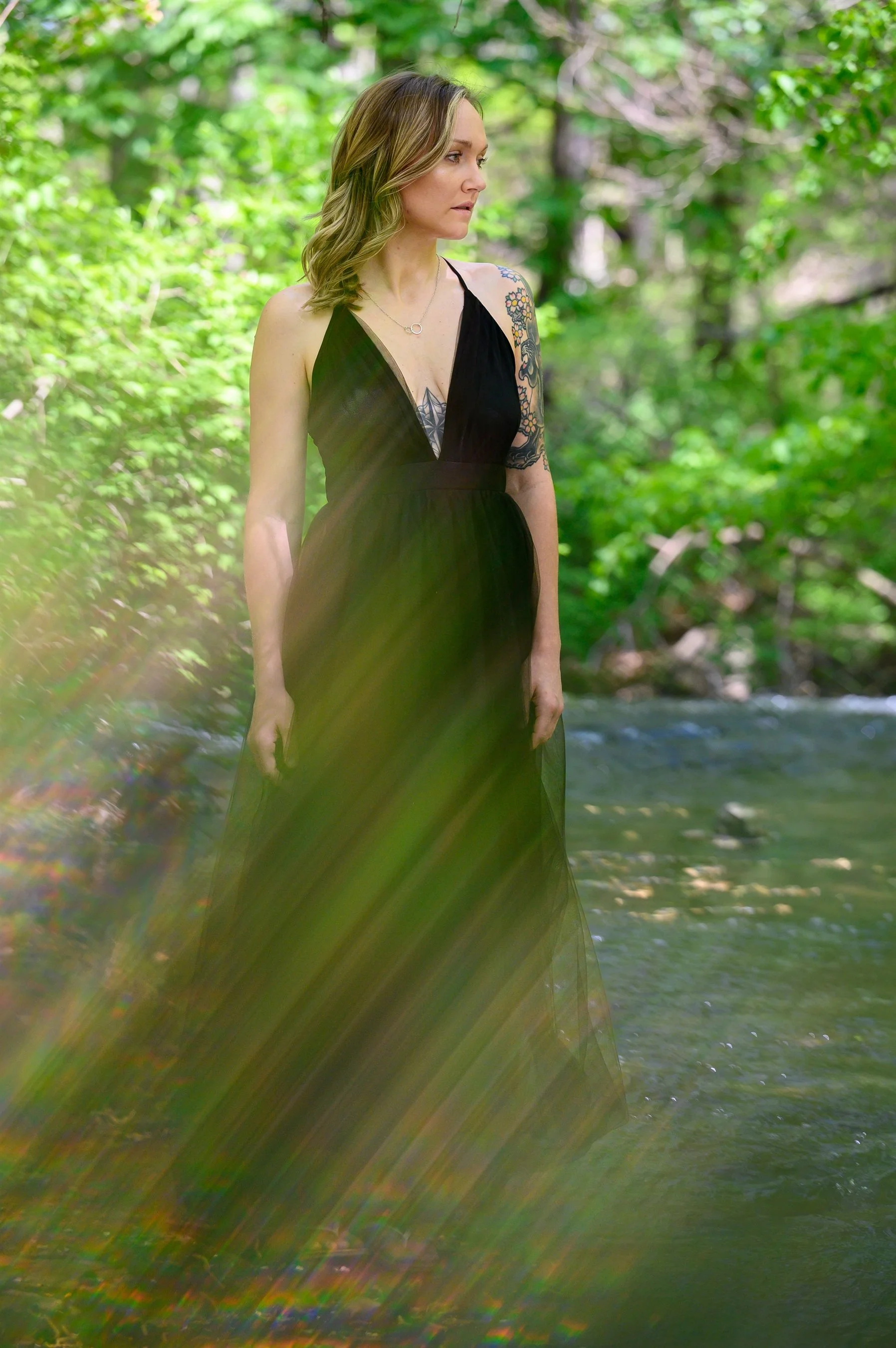 Woman in black dress with tattoos stands in a forest near a stream with sunlight creating a rainbow effect.
