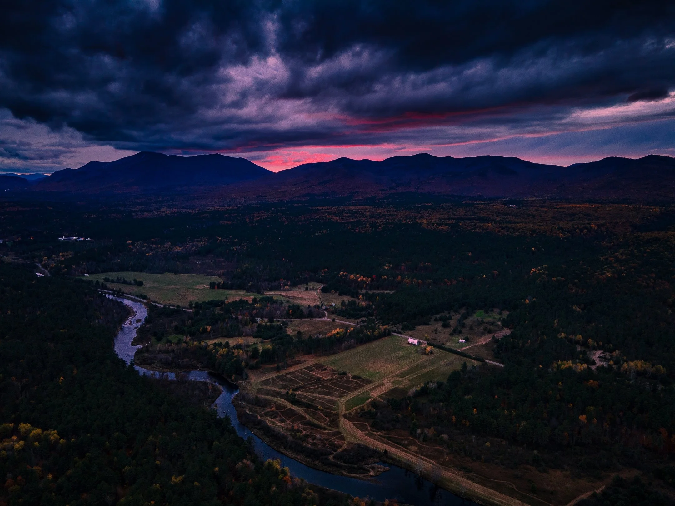 Aerial view of a landscape with a river flowing through a forest and fields, under a dramatic purple and blue sunset sky with mountains in the background.