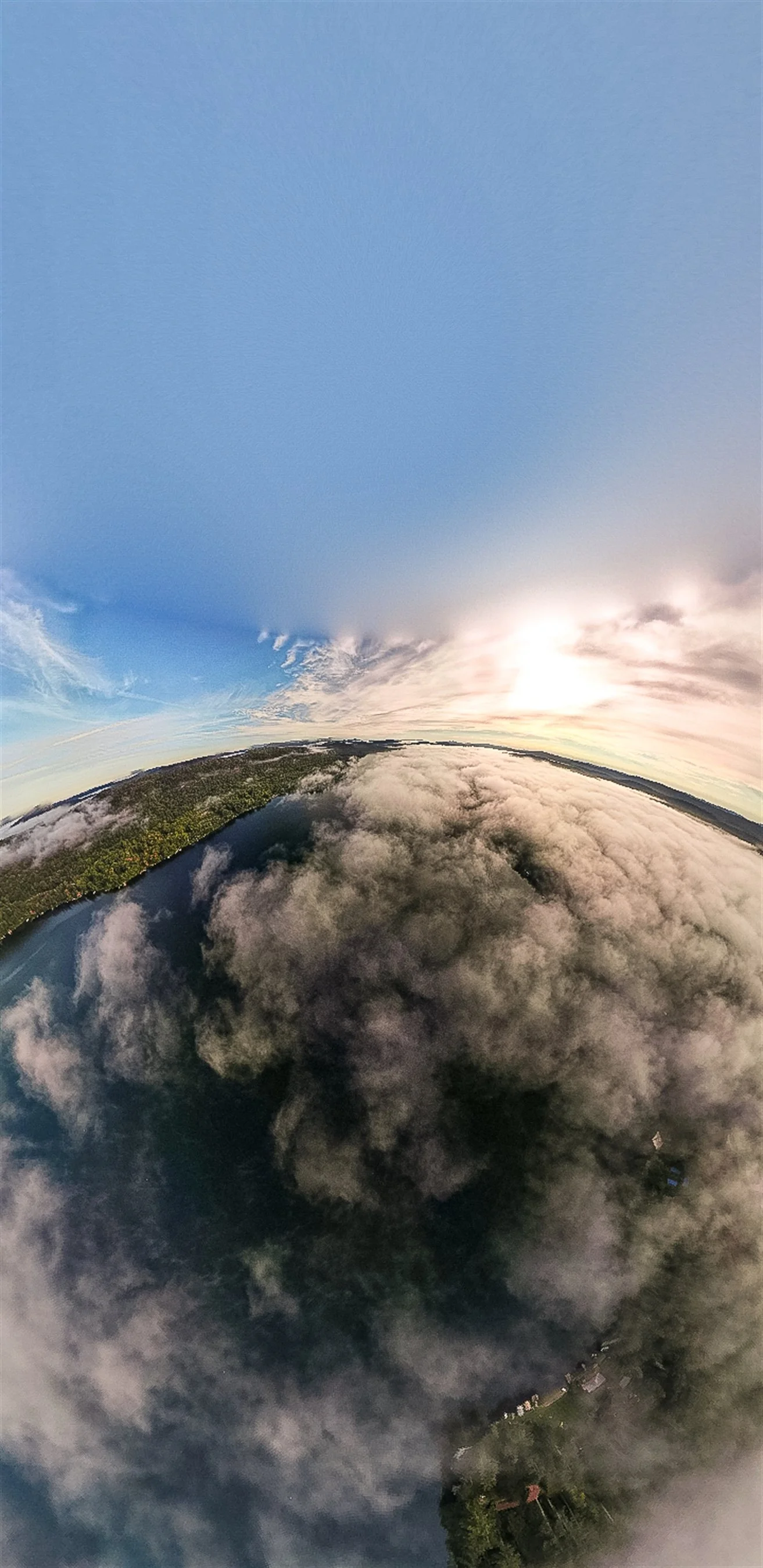 Aerial panoramic view of lush green forest and a body of water partially covered by fluffy clouds, with a clear blue sky above.