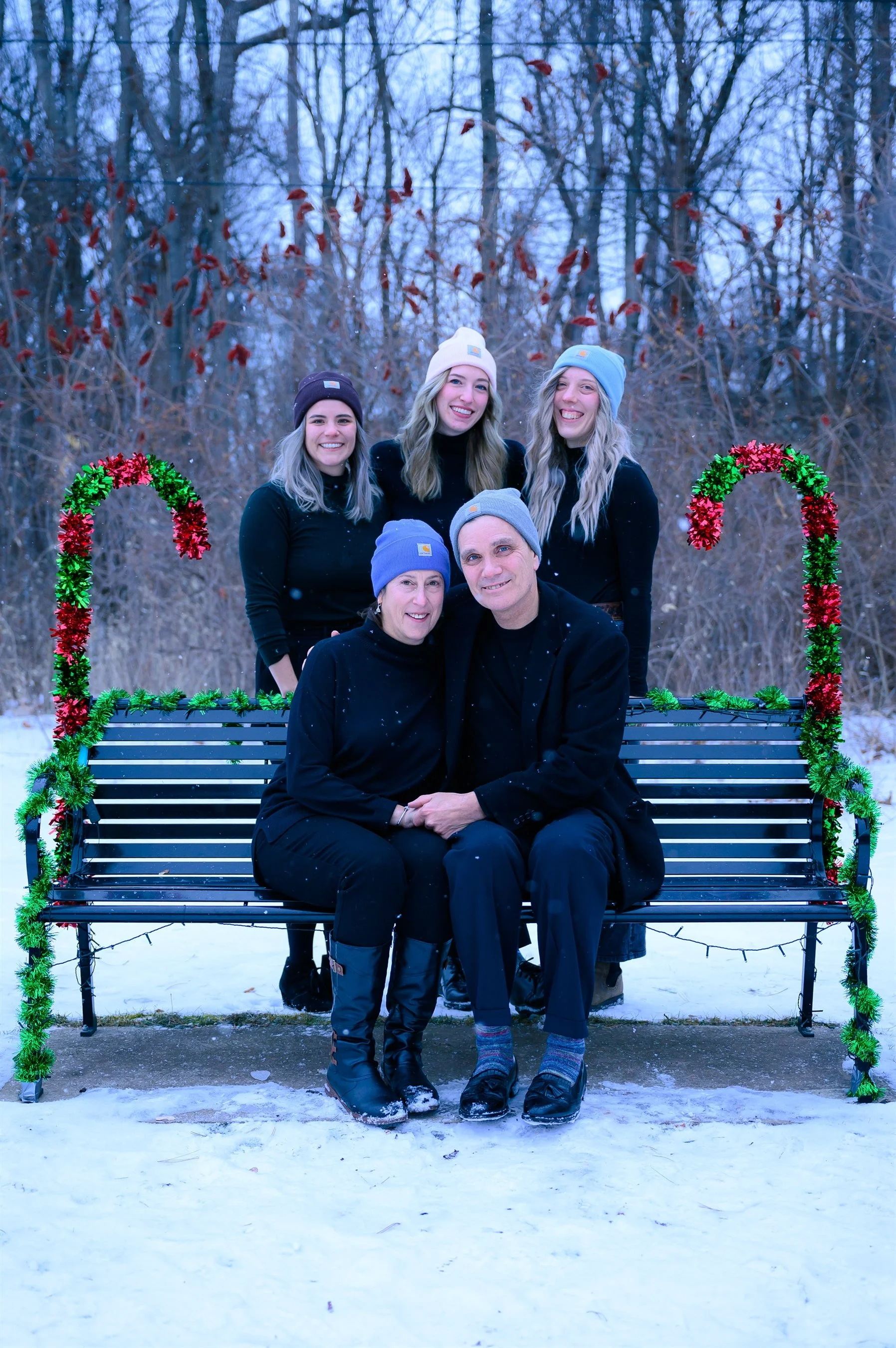 A family of five posed outdoors, sitting on a decorated bench with red and green tinsel. It's snowy, and they are all wearing beanies and black outfits. Two adults are seated in the front, and three young adults stand behind on the bench.