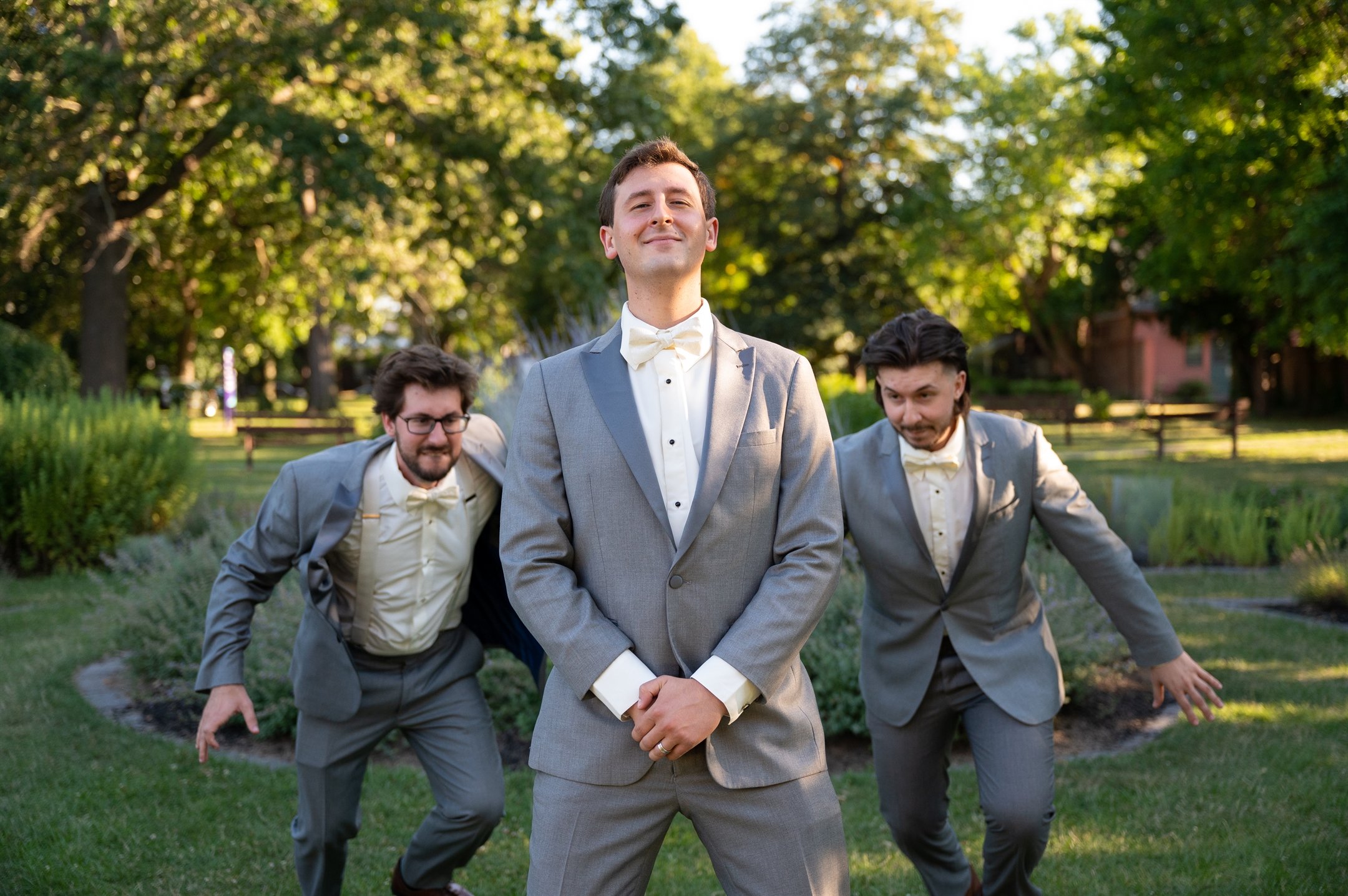 Three men in gray suits, one standing confidently in the center, while the other two playfully pose behind him in a garden setting.