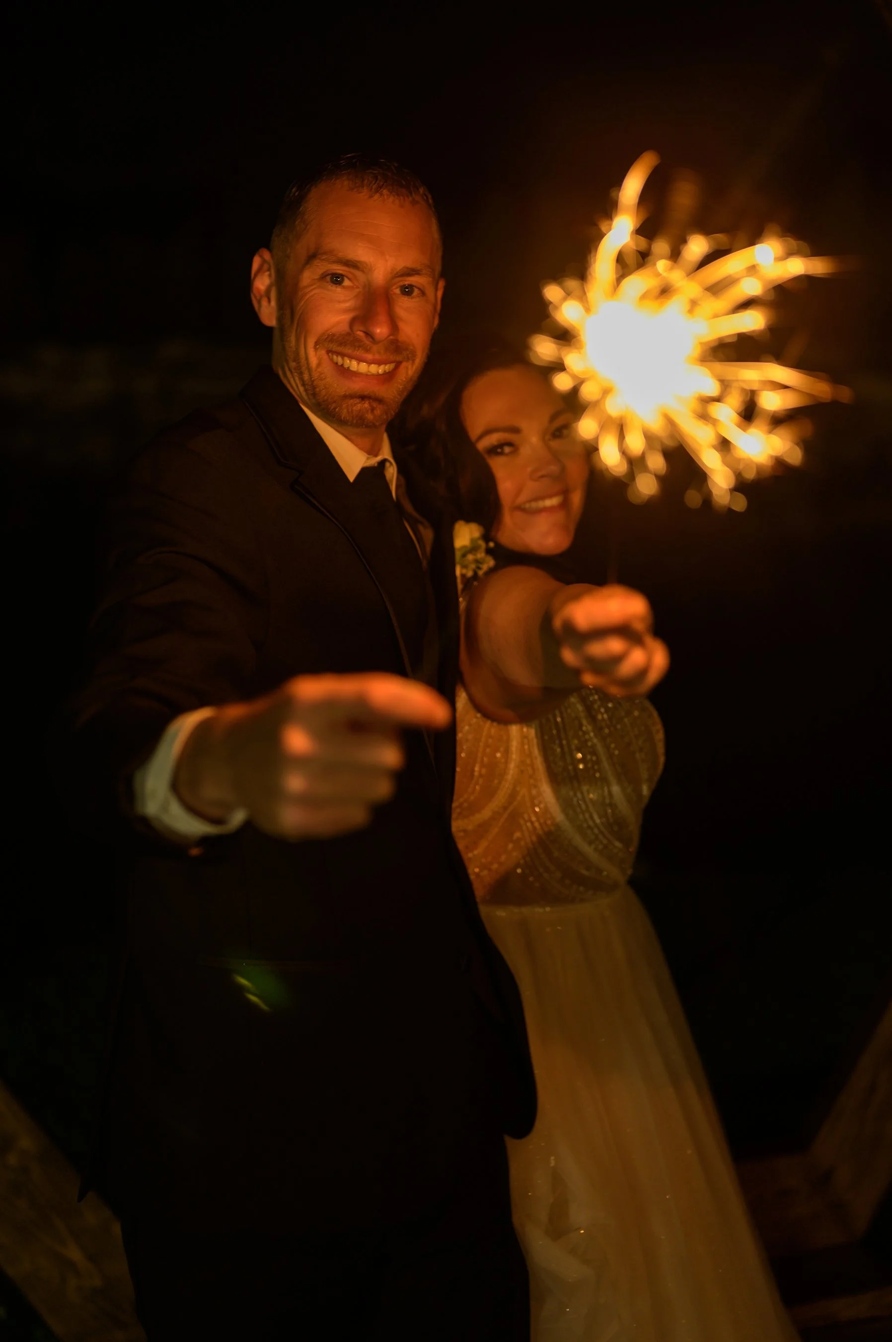 A couple dressed formally, holding a sparkler in the dark, smiling at the camera.