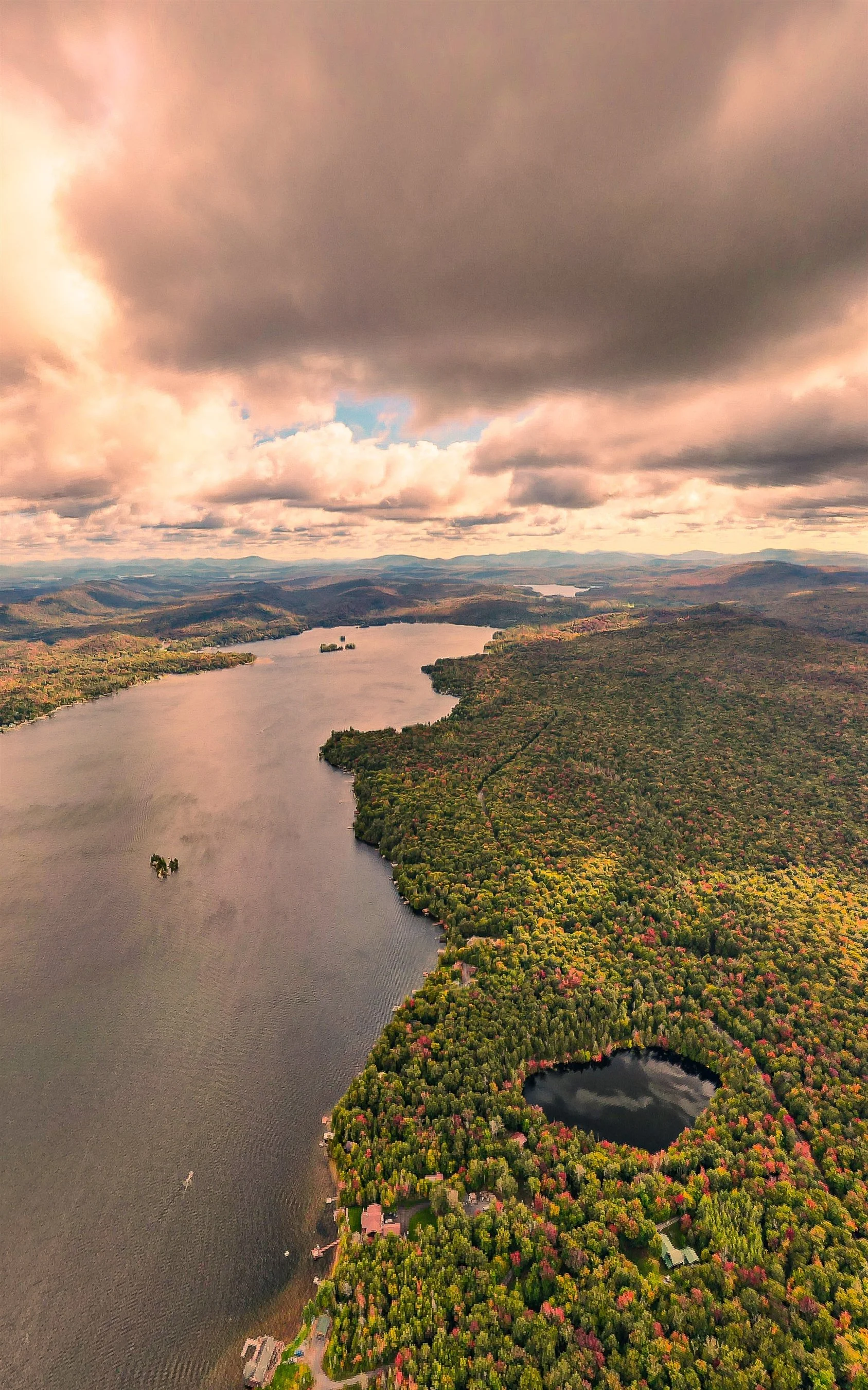 Aerial view of a large lake surrounded by dense forests and rolling hills with autumn foliage, under a dramatic cloudy sky.