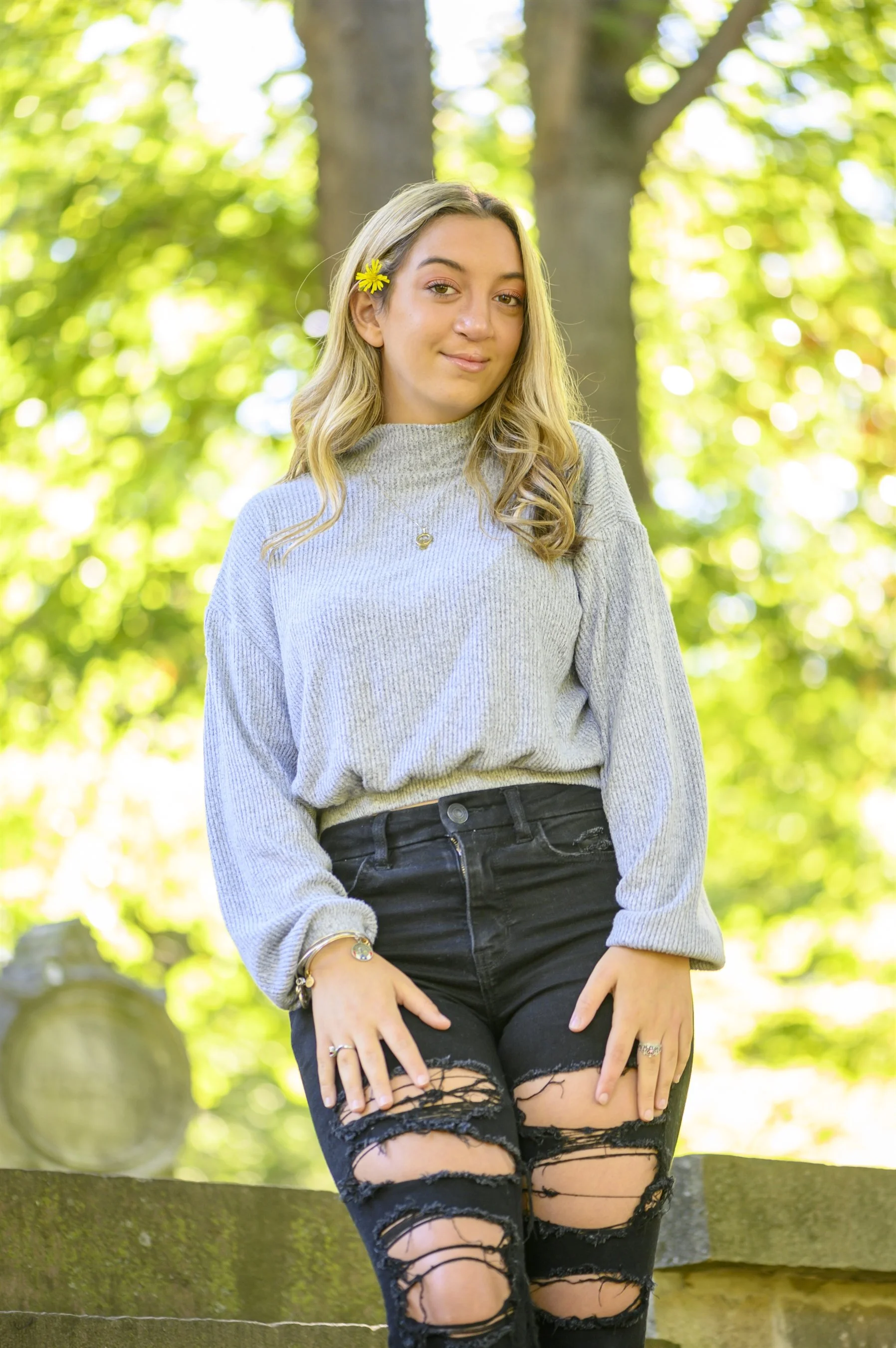 Young woman with long blonde hair, wearing a gray sweater and ripped black jeans, standing outdoors with trees in the background; a yellow flower is tucked behind her ear.