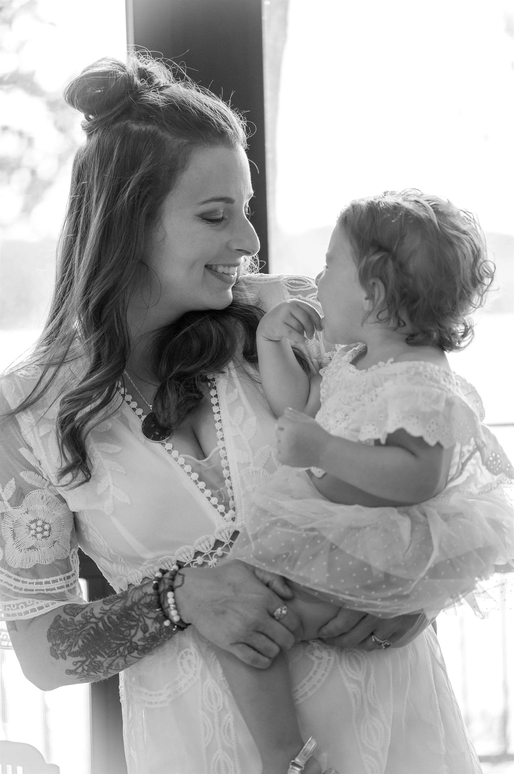 Woman holding a toddler and smiling, both wearing light-colored outfits, indoors.