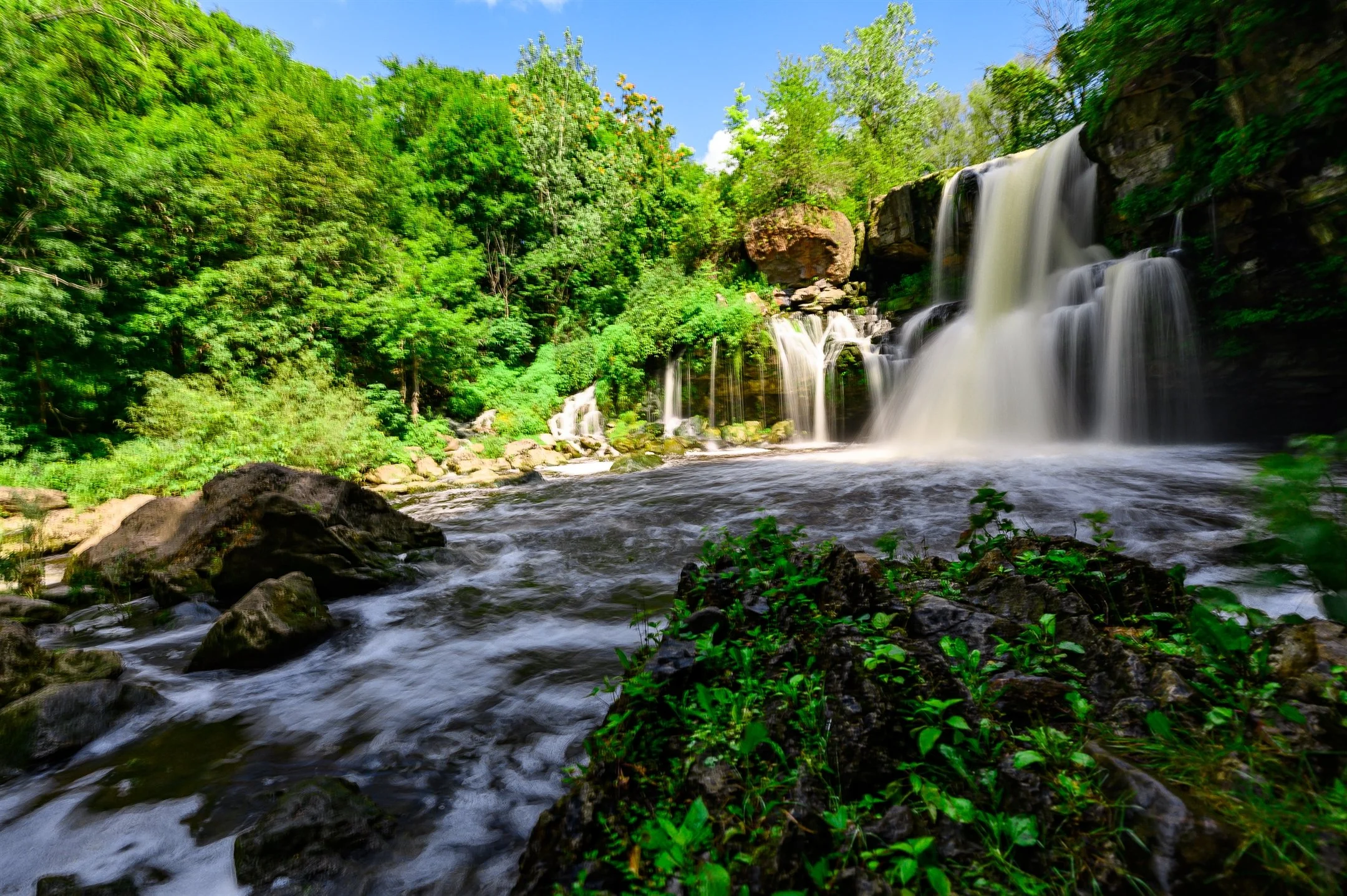 Waterfall surrounded by lush green forest, with rocks and flowing water in the foreground under a clear blue sky.