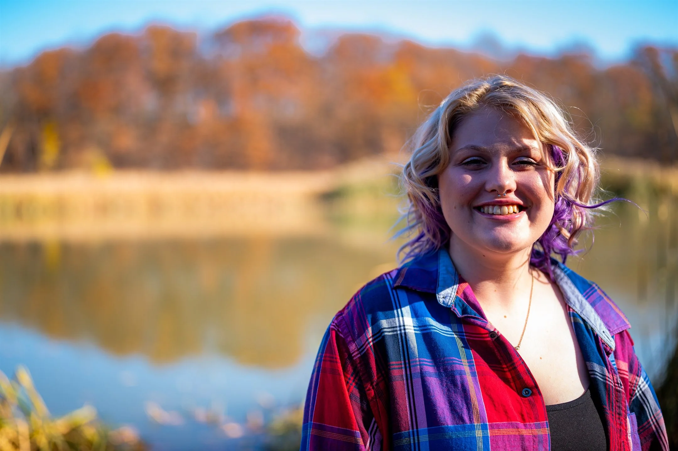 Smiling woman with blonde and purple hair in plaid shirt outdoors near a lake with autumn trees.