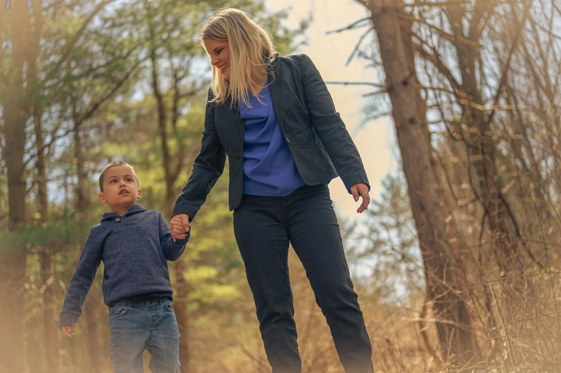 A woman and child holding hands, walking through a forest with sunlight filtering through the trees.