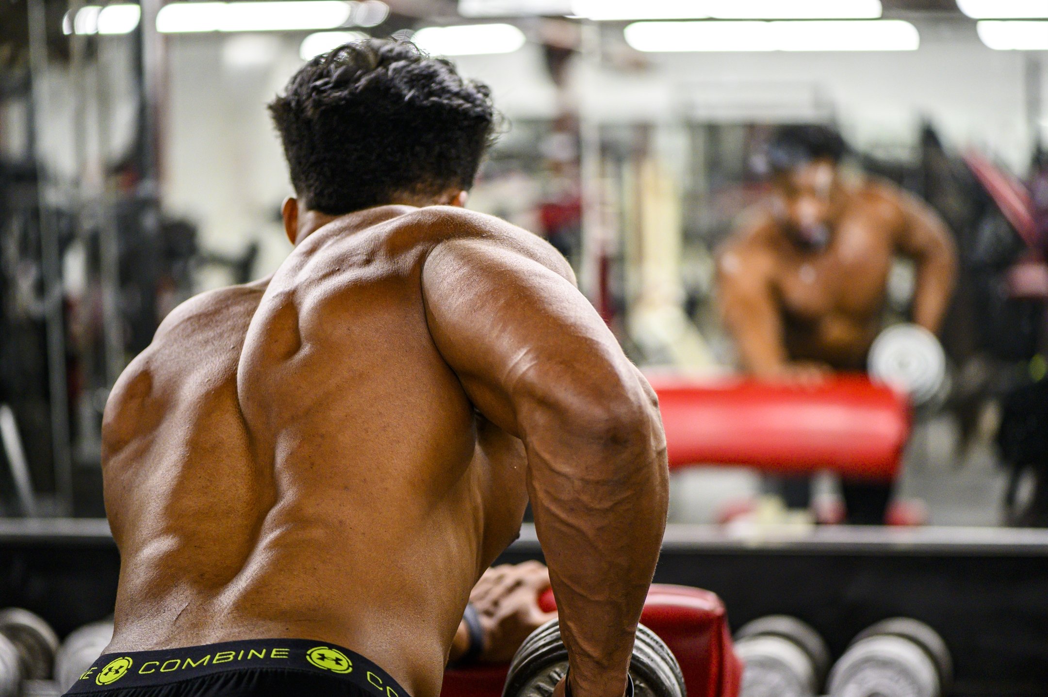 A muscular person with a bare back holding a dumbbell, exercising in a gym, with their reflection visible in the mirror.
