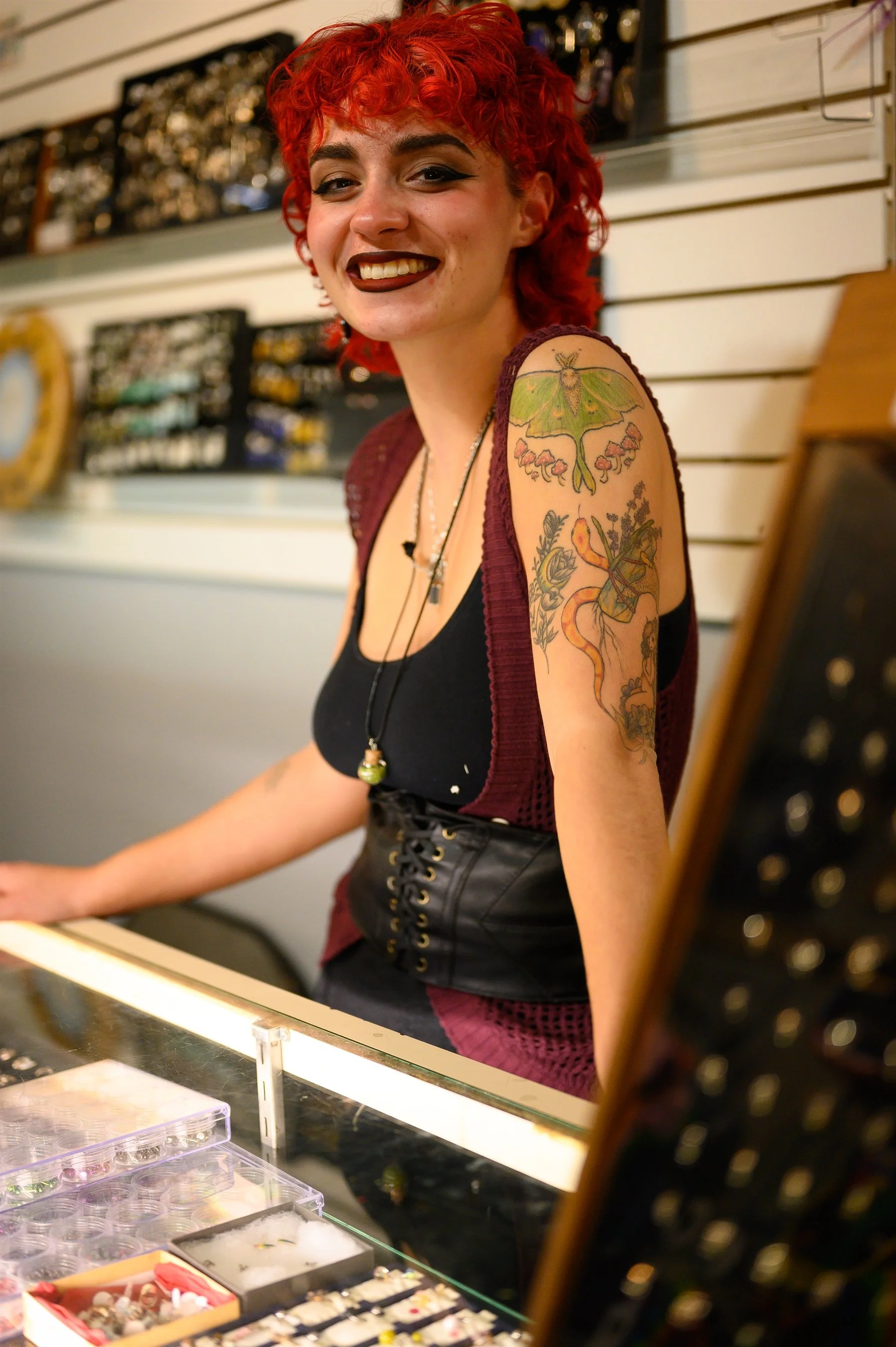 Smiling person with red curly hair and tattoos, standing behind a jewelry display case in a store.