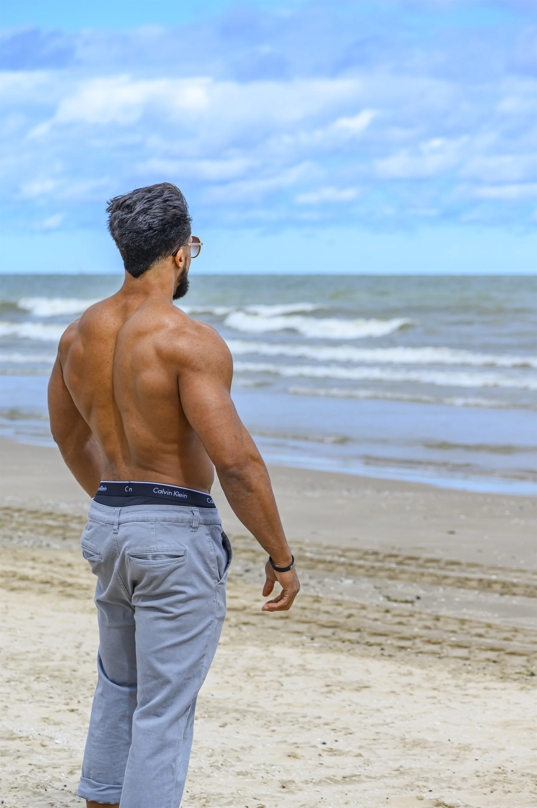 Shirtless man standing on a beach facing the ocean, with sunglasses and gray pants.