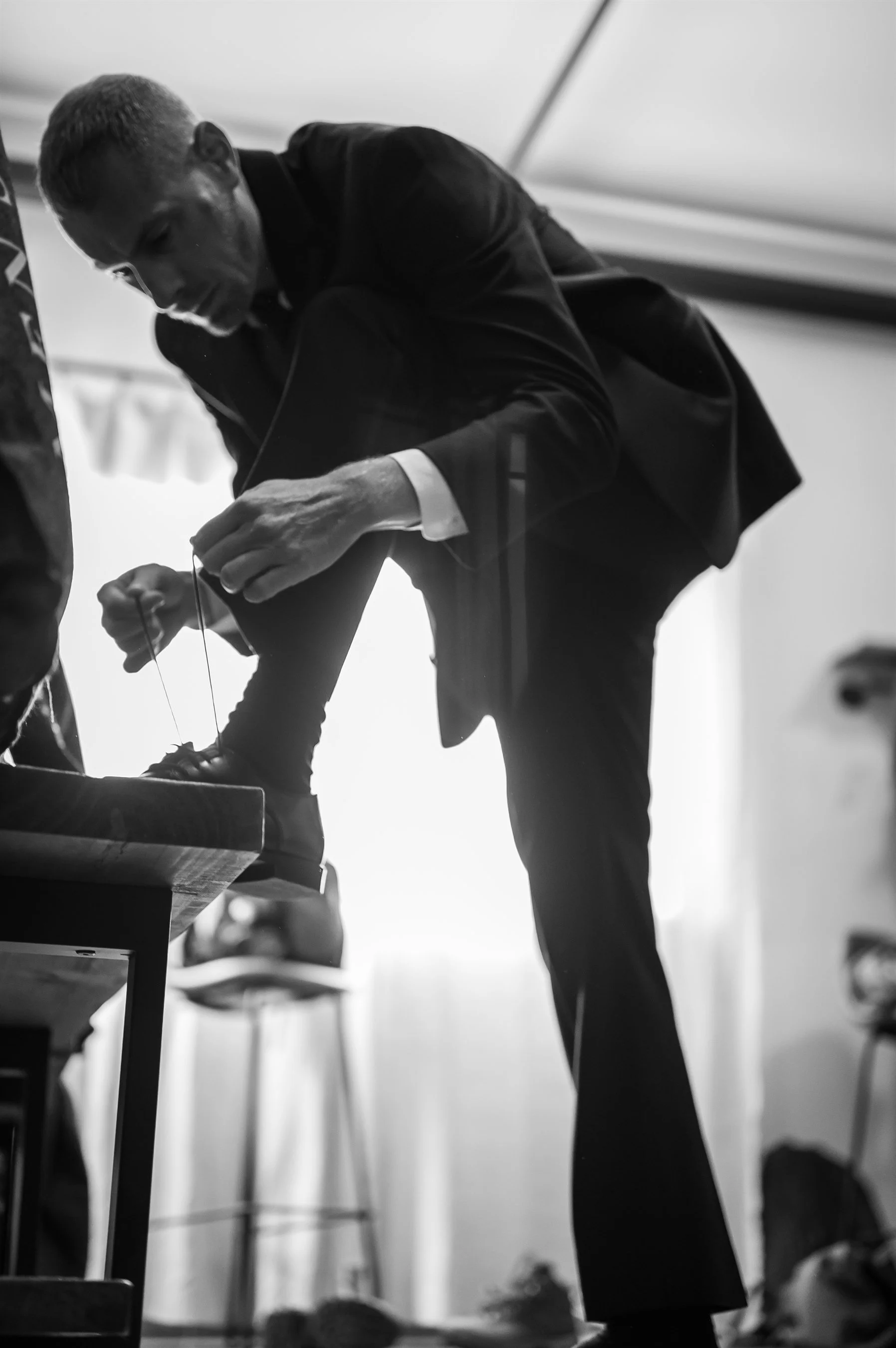 Black and white photo of a man in a suit tying his shoe, kneeling with one leg on a chair.