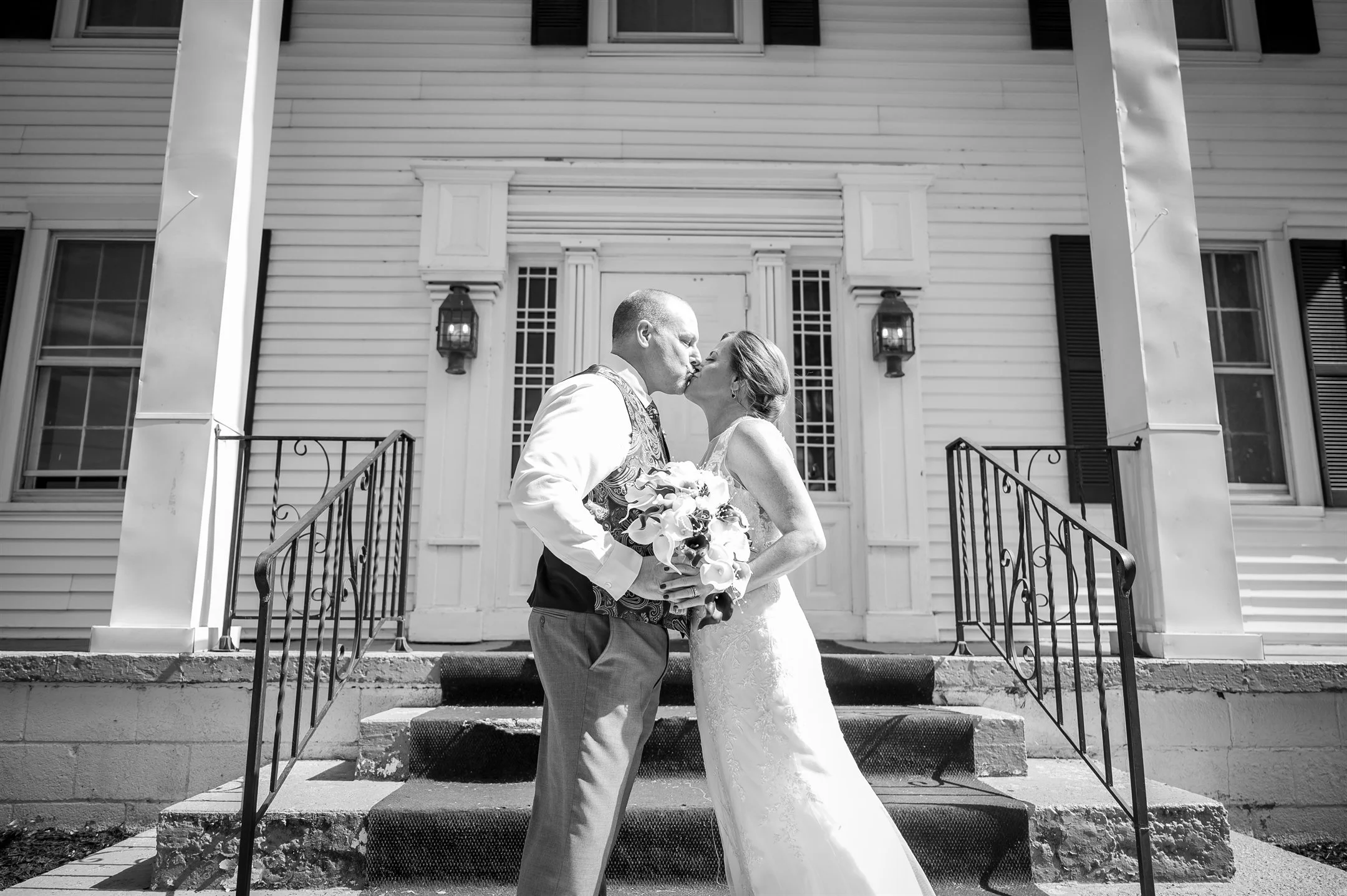 Bride and groom kissing on outdoor steps, holding flowers, in front of a white building with pillars and lanterns.