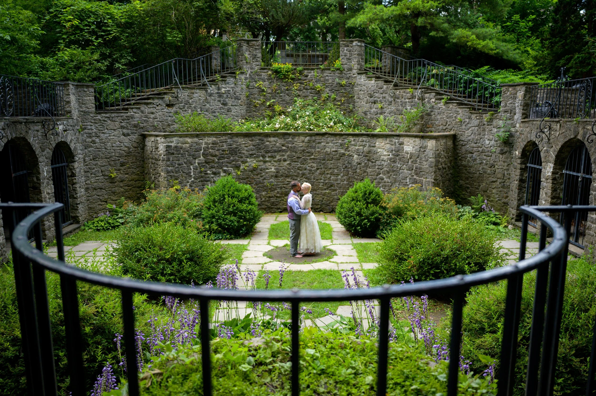 A couple embracing in a lush garden surrounded by stone walls and greenery, with a wrought-iron gate in the foreground.