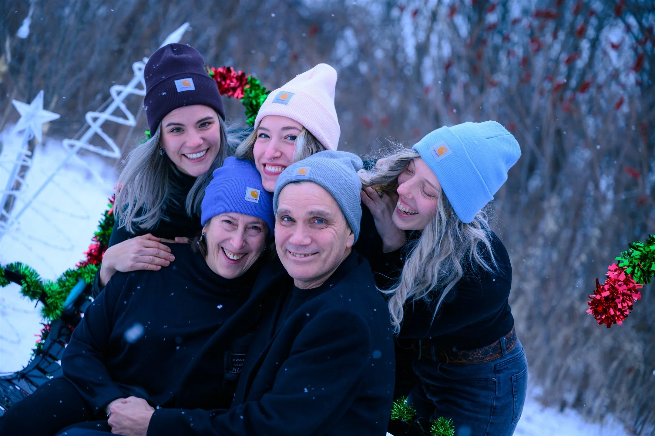 A family of five wearing winter hats and black clothing sitting outdoors in a snowy setting. They are smiling and hugging, surrounded by holiday decorations.