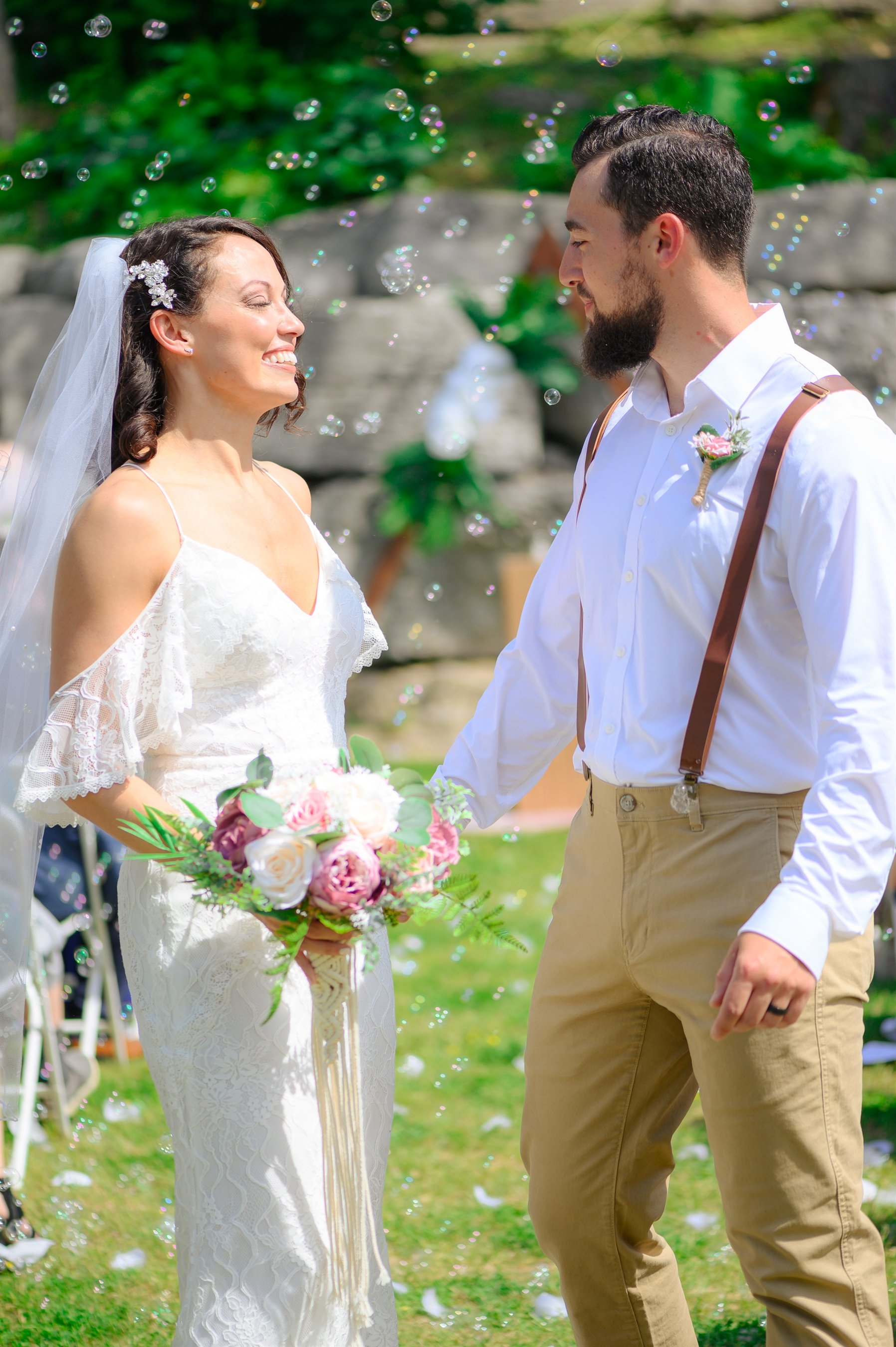 Bride and groom smiling at each other outdoors, wearing wedding attire, surrounded by bubbles. The bride holds a bouquet of flowers.