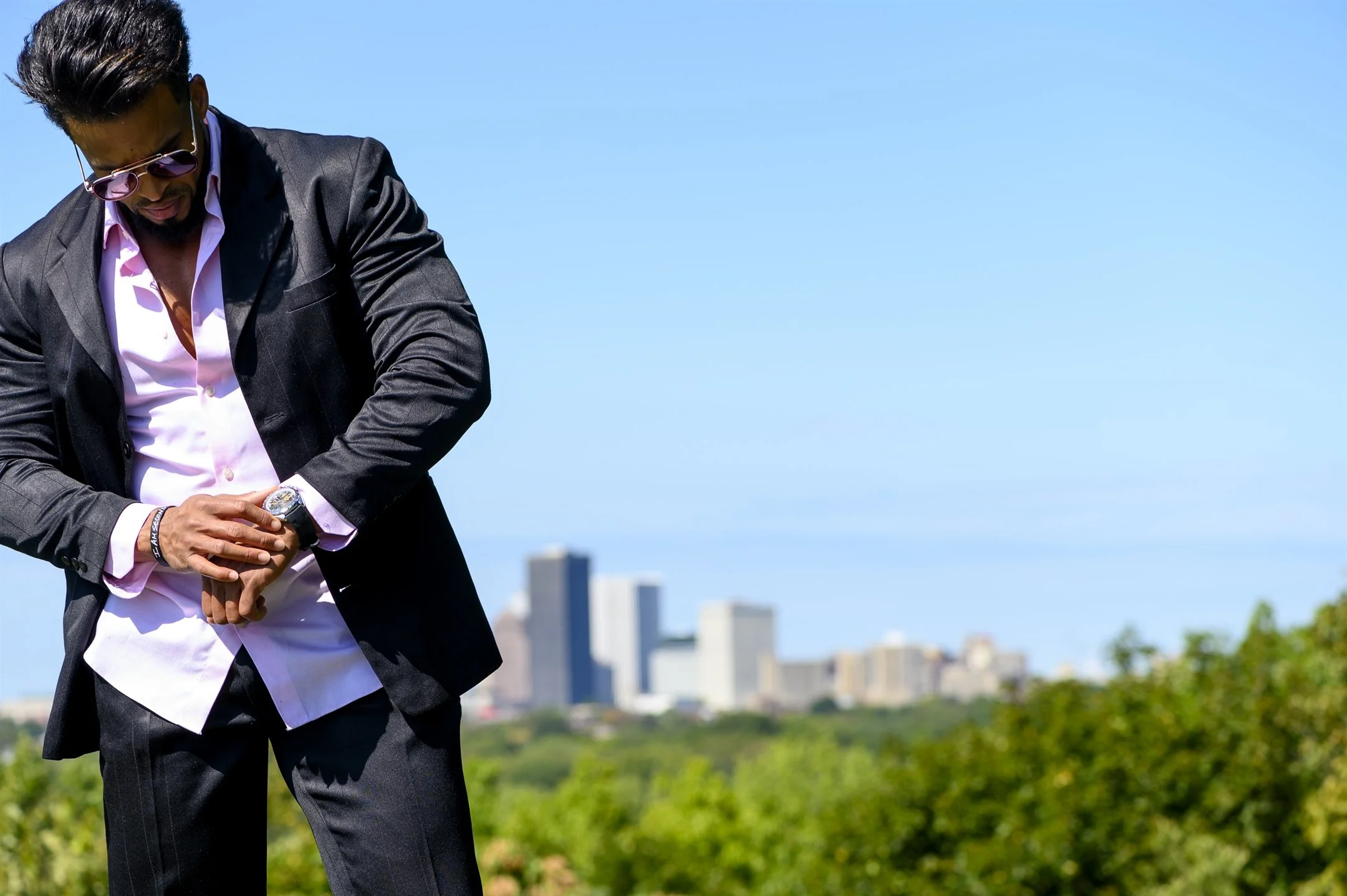 Man in sunglasses and a suit checking his watch with a city skyline in the background.