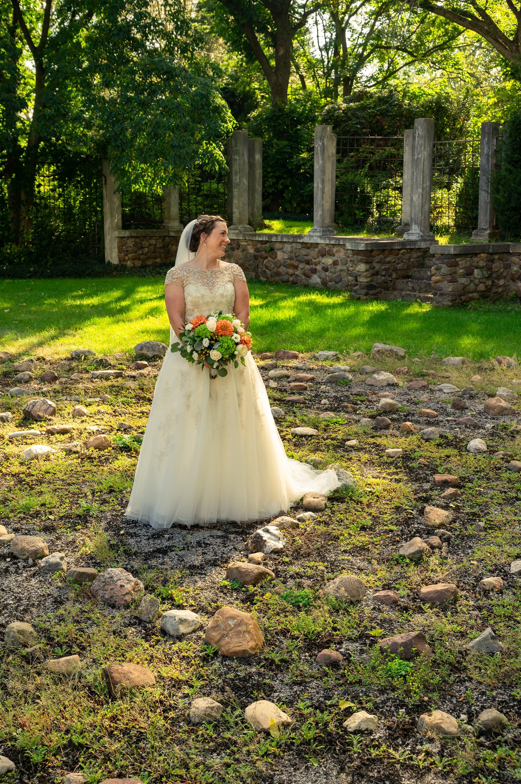 Bride in white dress holding a bouquet, standing in a garden with stone circle pathway, surrounded by trees and stone pillars.