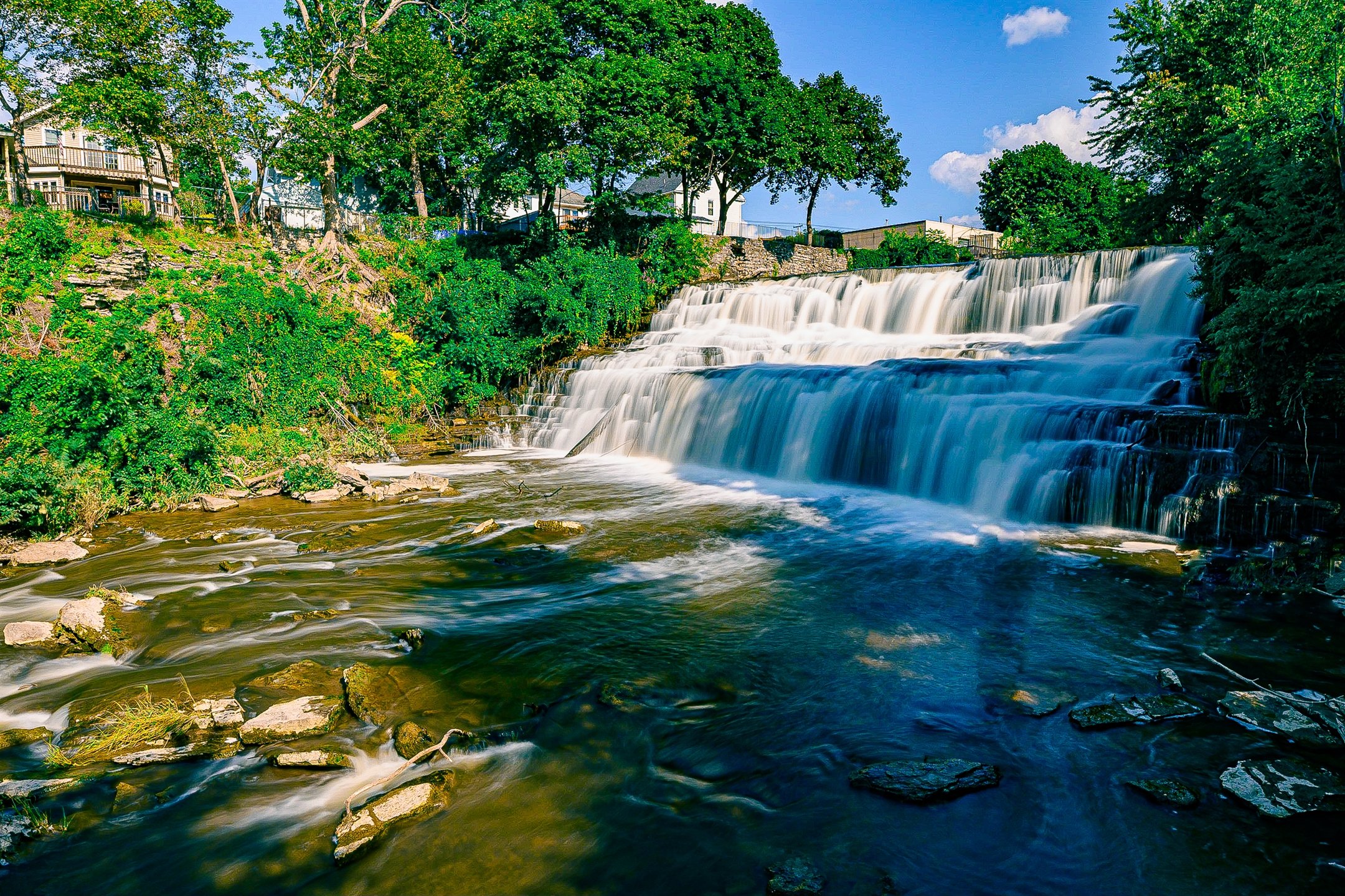 A waterfall flowing over rocks with trees and houses in the background on a sunny day.