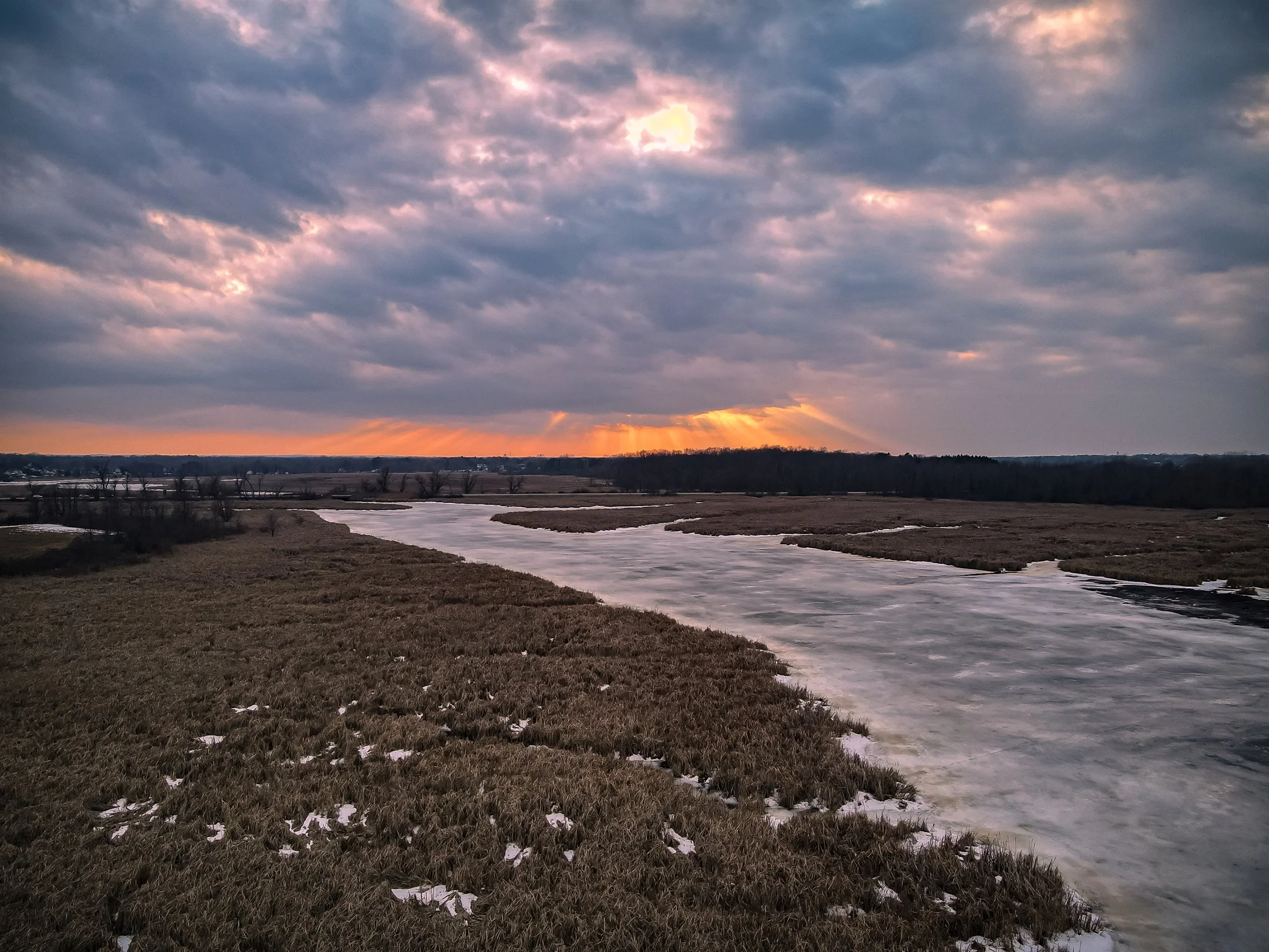 A picturesque winter landscape featuring a frozen river winding through a snow-dusted field, with scattered patches of brown grass. The sky is overcast, with dramatic clouds and sun rays peeking through, casting a warm glow on the horizon.