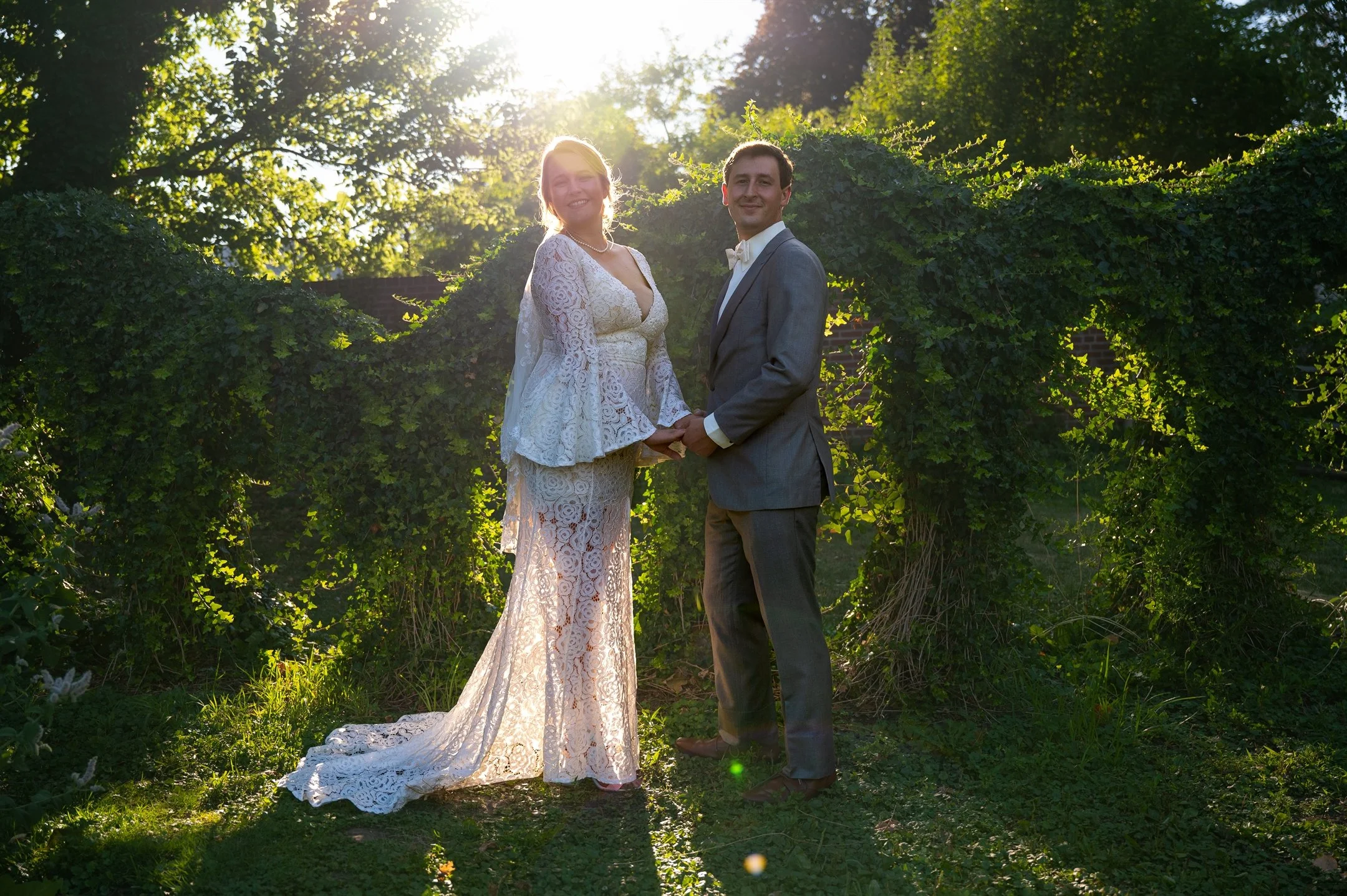 A couple holding hands in a garden, with the woman in a lace wedding gown and the man in a gray suit, standing in front of sunlit greenery covered with ivy.