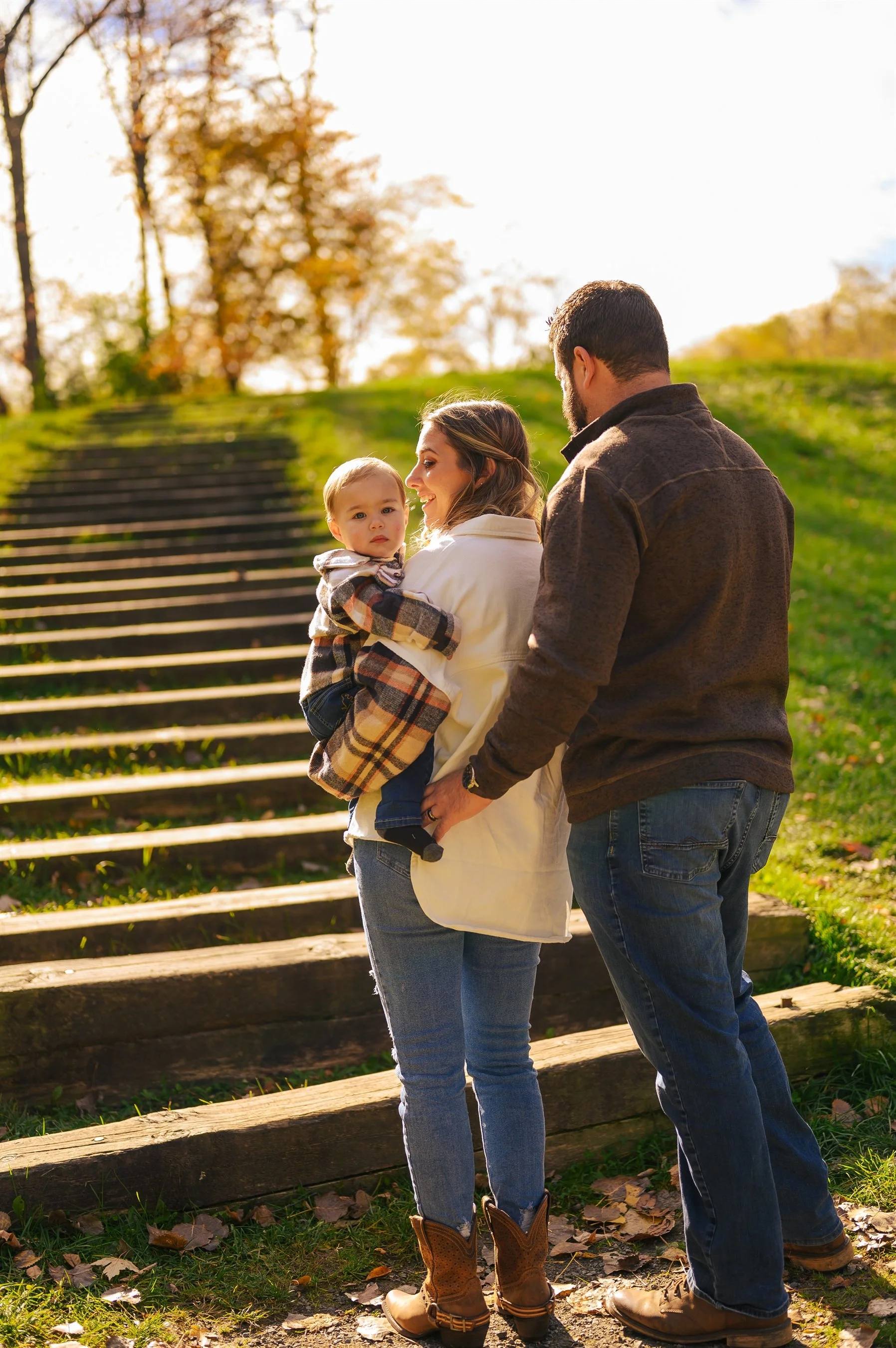 A family standing outdoors near steps, with a woman holding a baby, and a man standing beside her. The background features trees and sunlight.