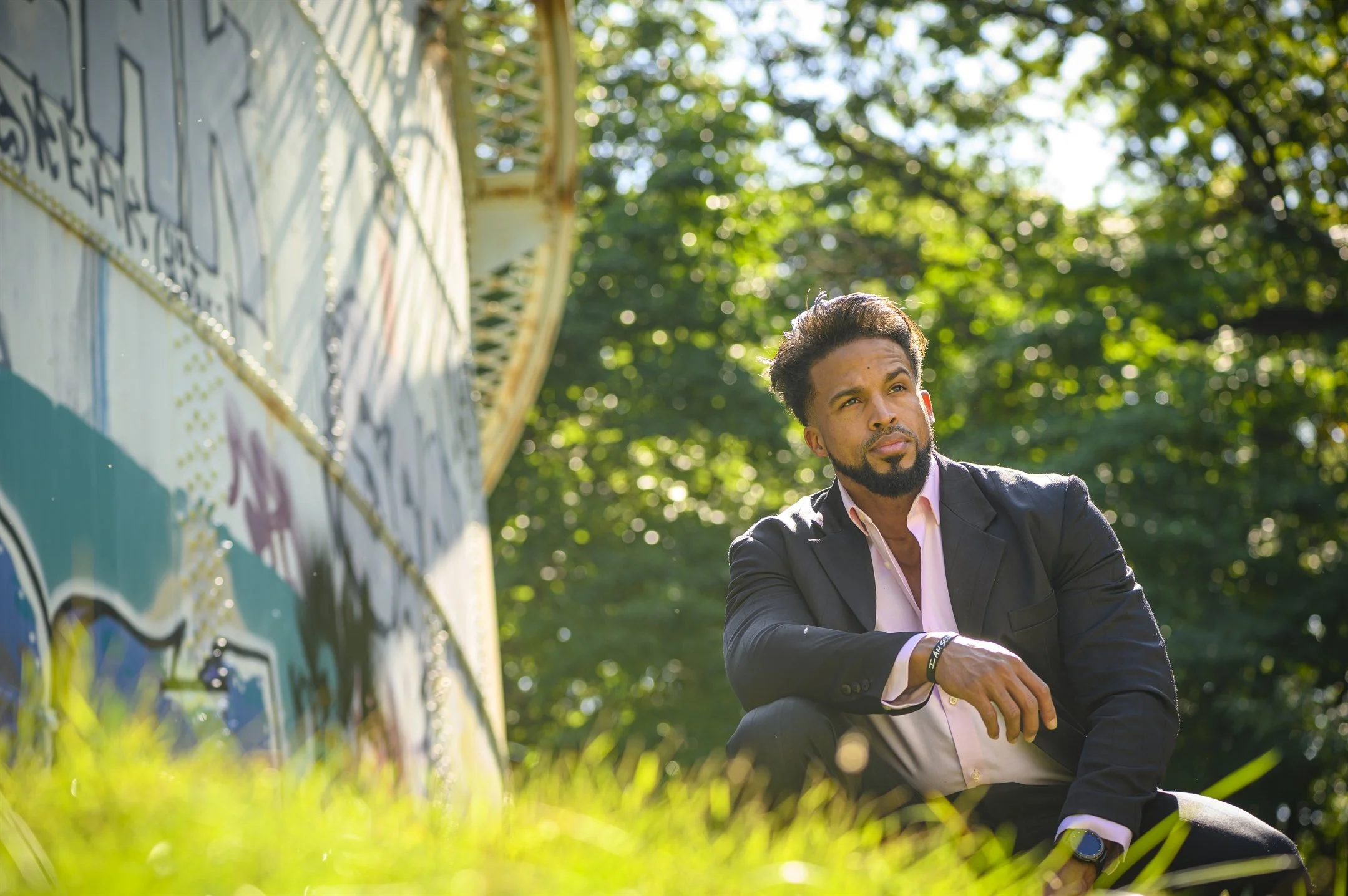 Man in a suit sitting outdoors near a graffiti-covered structure, surrounded by trees and sunlight.