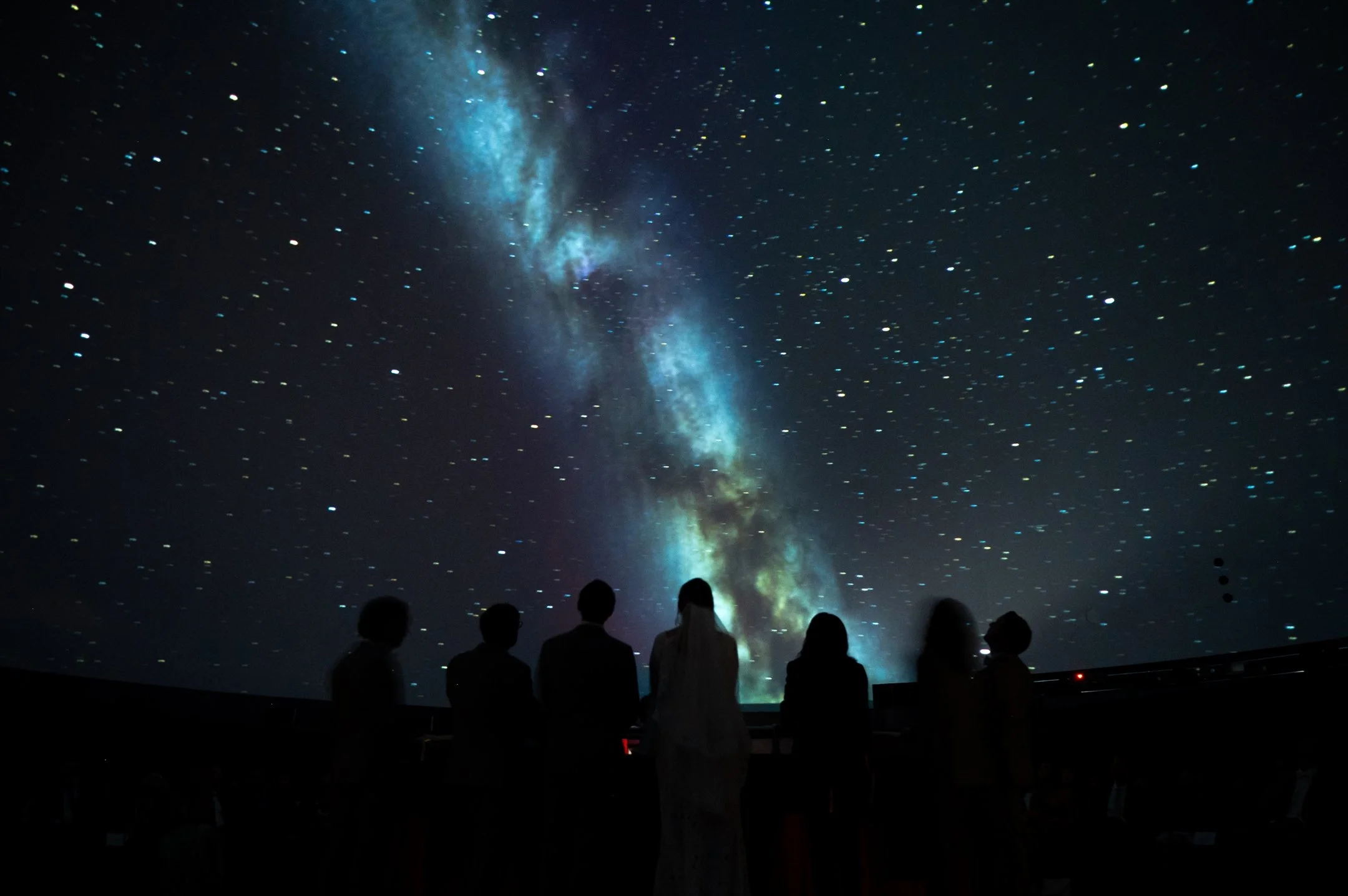 Silhouetted people standing in front of a starry sky projection, featuring the Milky Way.