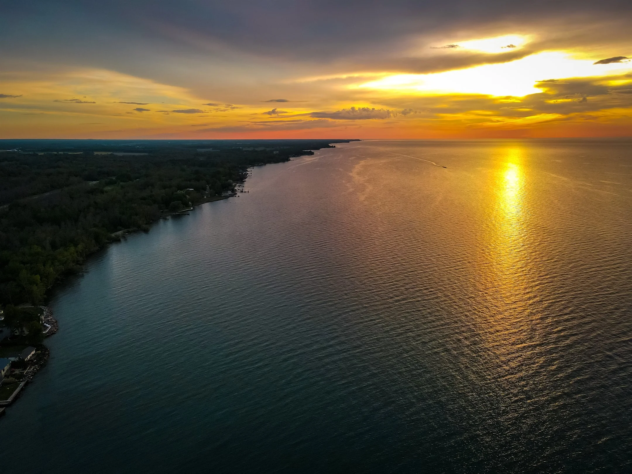 Aerial view of a calm lake with surrounding forested shore at sunset, reflecting vibrant orange and yellow hues on the water.