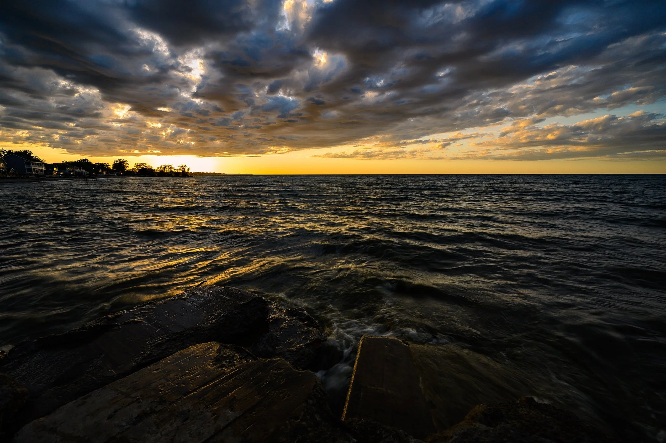 Dramatic sunset over a body of water with dark clouds and golden reflections on the waves. Coastal houses and trees are silhouetted on the horizon.