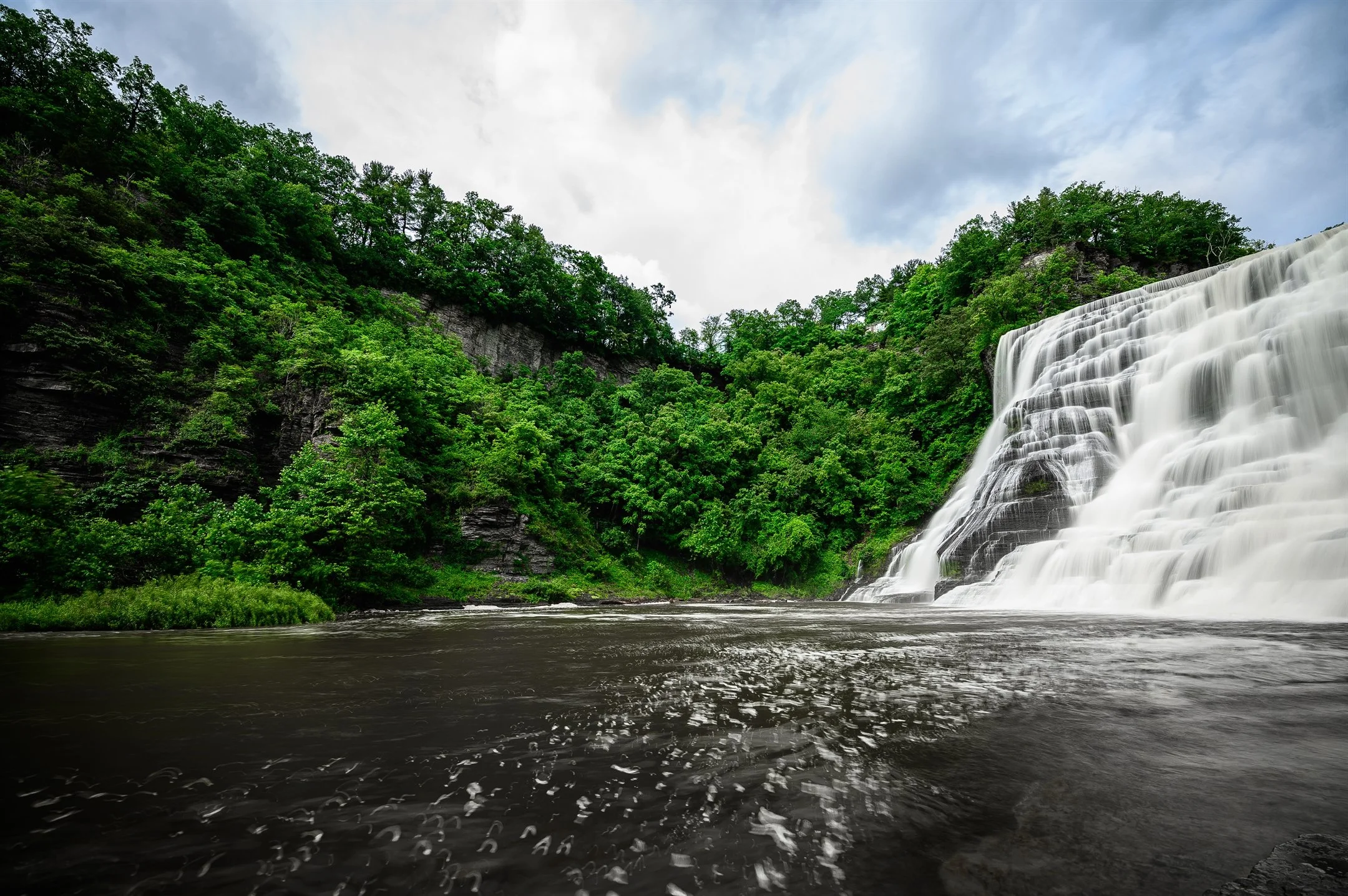 A waterfall cascading down a rocky cliffside surrounded by dense green forest, with a river flowing beneath under a cloudy sky.