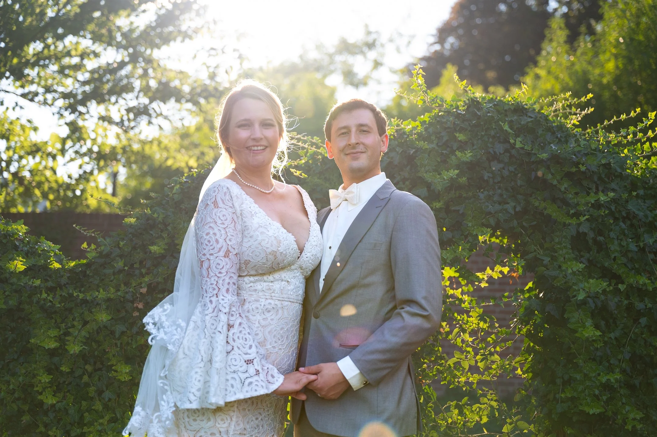 Bride and groom in outdoor garden, holding hands, bride in lace dress, groom in gray suit.