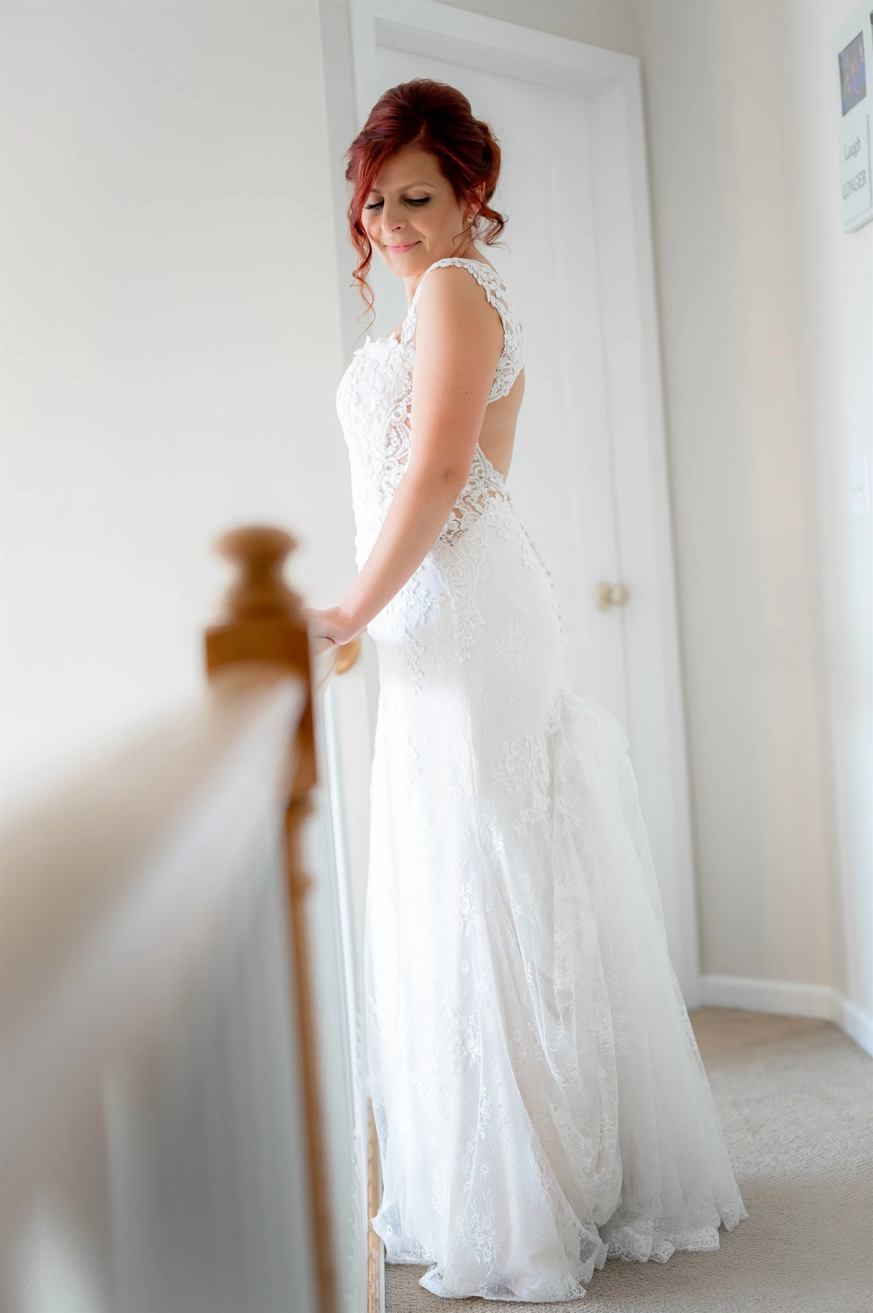 Bride in lace wedding dress standing indoors near staircase railing, looking down and touching the banister.