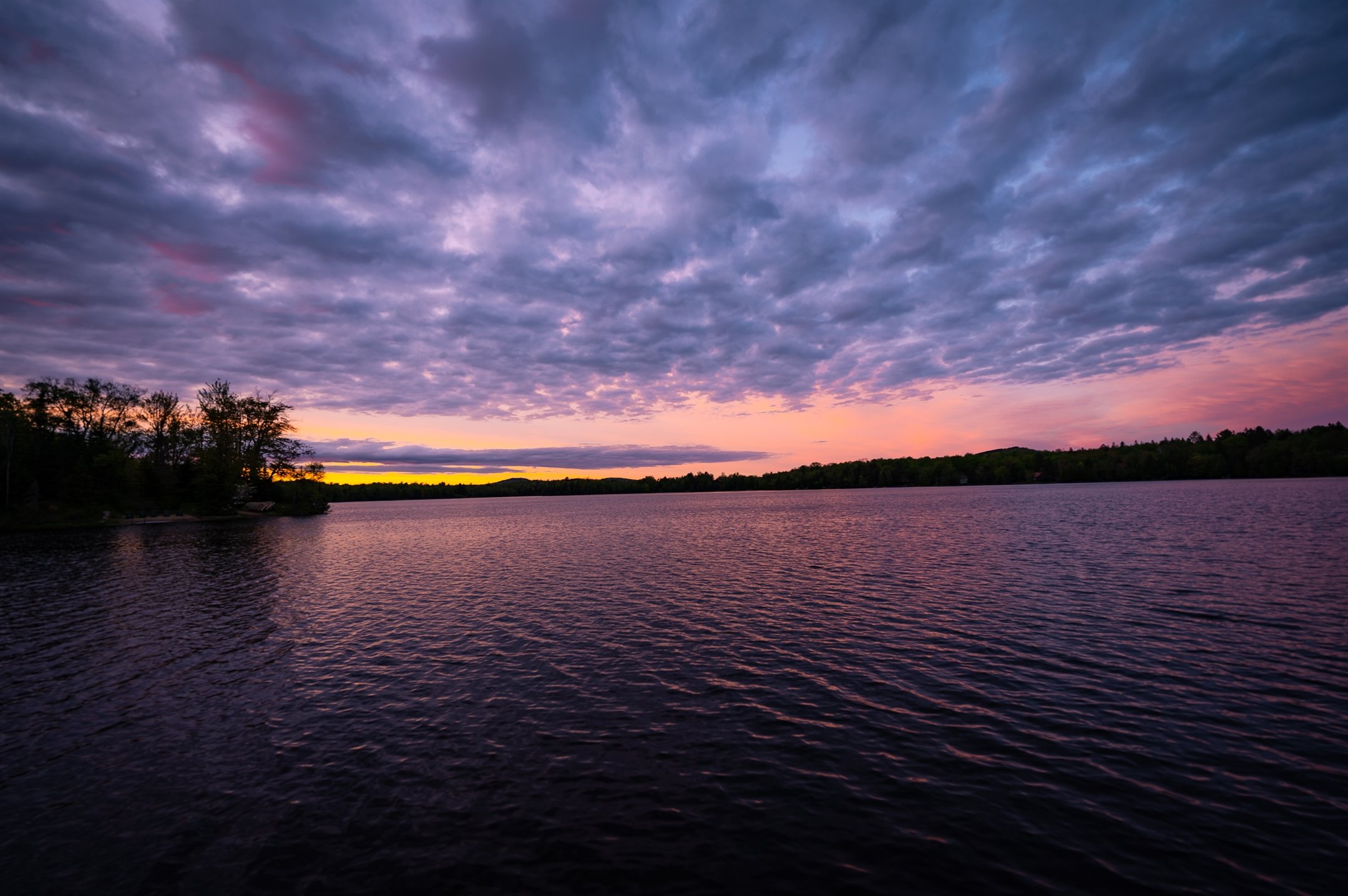 Lake at sunset with colorful sky, clouds, and distant trees on the shore.