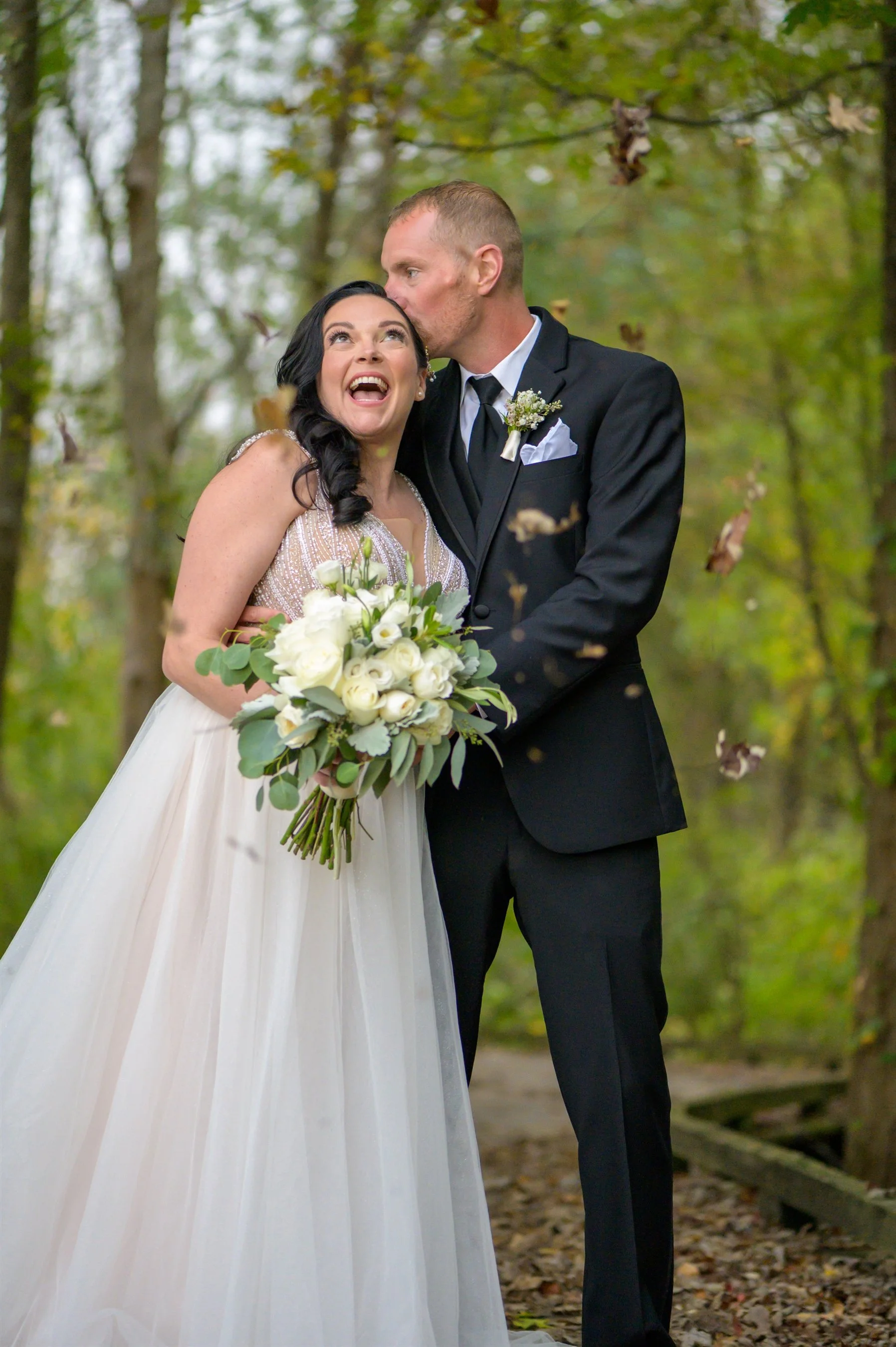 A bride and groom in a forest, the bride holding a bouquet with white flowers and greenery. The groom kisses the bride's forehead. Both appear happy, with fallen leaves in the background.