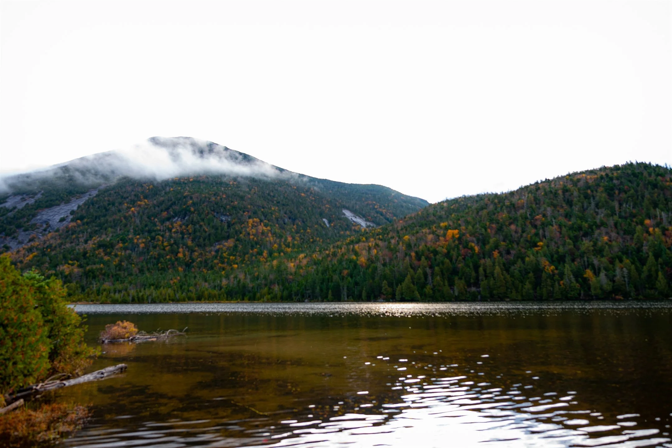 Scenic view of a lake with calm water, surrounded by forested hills with fall foliage, and low-hanging clouds over the hills.