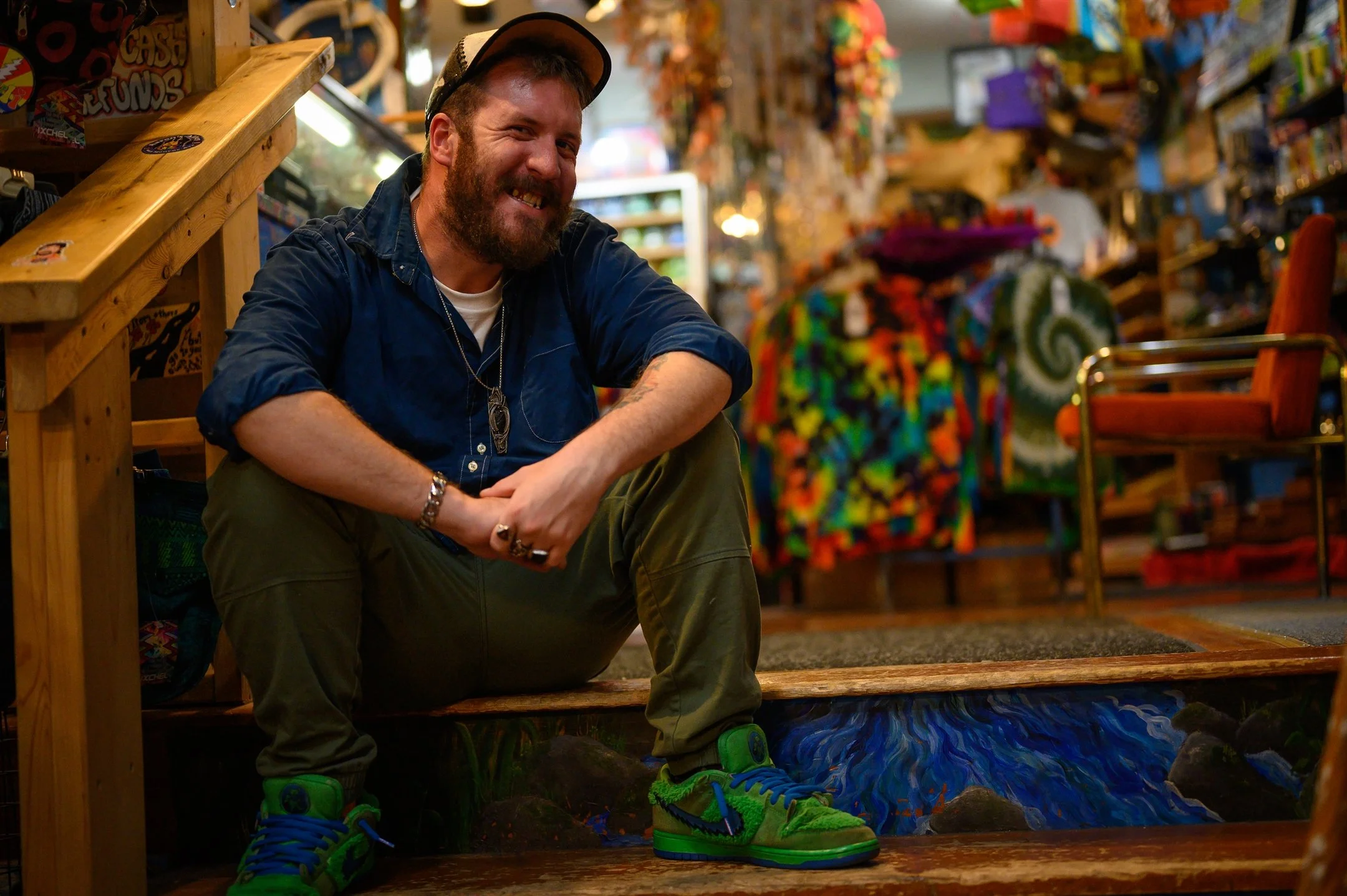 Man sitting on steps in colorful shop, wearing green sneakers, a blue shirt, and a cap, with tie-dye clothing visible in the background.