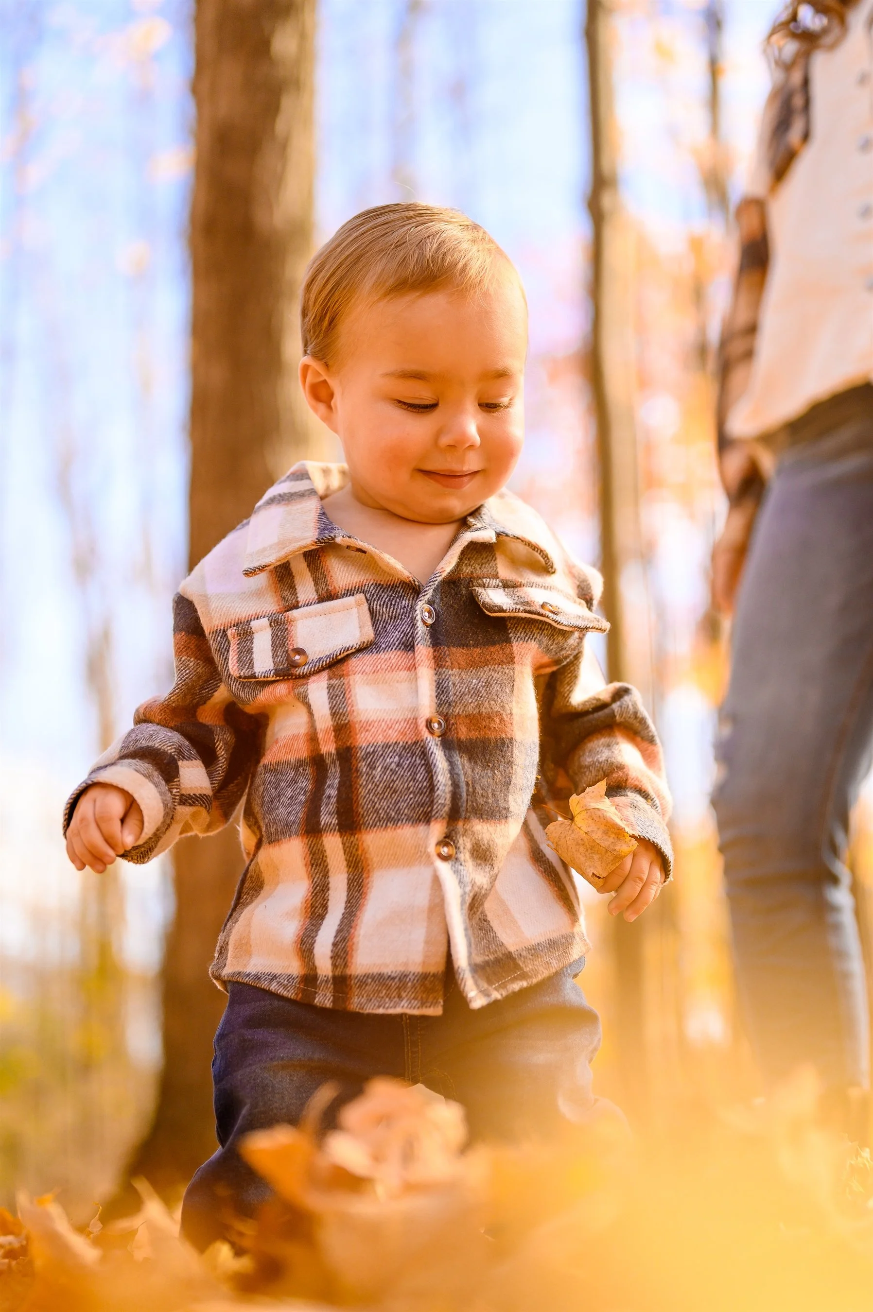 A young child wearing a plaid jacket and jeans walking through autumn leaves in a forest. Another person is partially visible in the background.